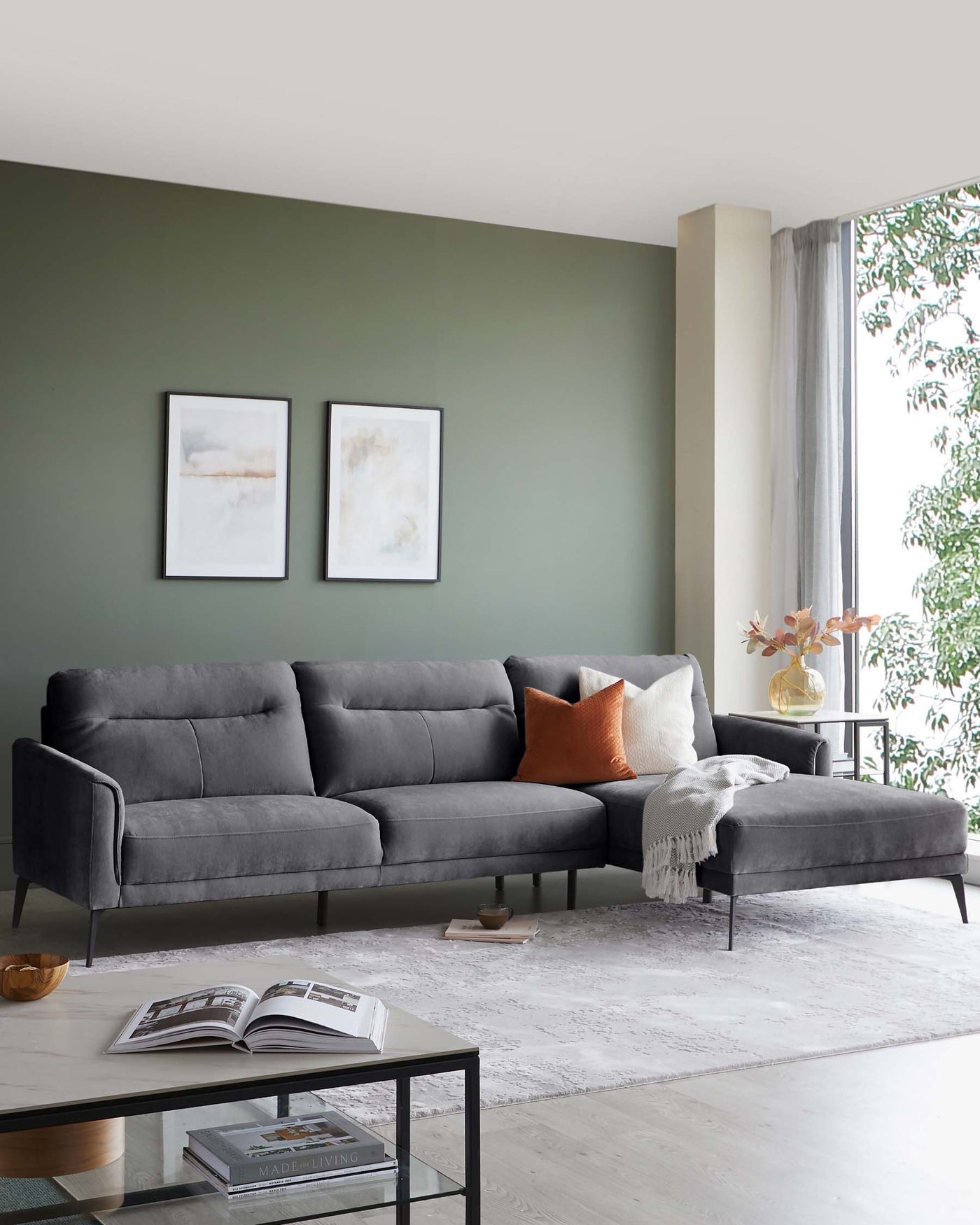 Elegant modern living room with a charcoal grey sectional sofa featuring clean lines, tufted back cushions, and a matching chaise lounge. A sleek black metal-framed coffee table with a glass top and books rests on a textured white area rug.