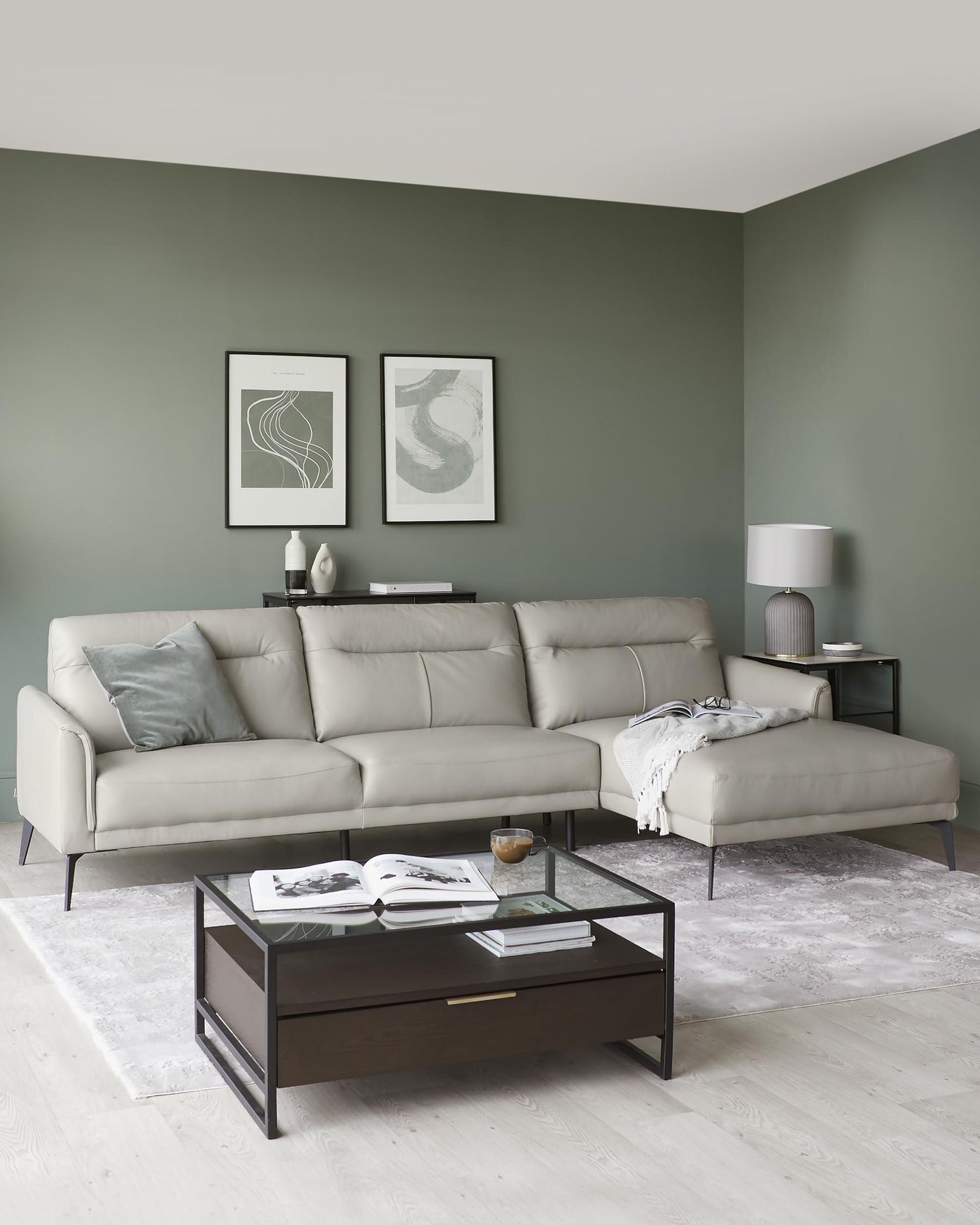 A contemporary light grey sectional sofa with a chaise lounge on its right end, featuring minimalist metal legs, complemented by a dark wood coffee table with a glass top and metal frame, resting on a soft textured area rug. A sleek, dark-finished side table with a simple table lamp is also present beside the sofa.