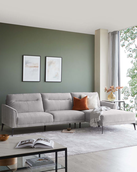 Modern light grey sectional sofa with tufted back cushions and a chaise lounge extension, accented with contrasting orange and white throw pillows and a light grey throw blanket. The black minimalist coffee table with glass top stands in front, beside which lies a hardcover book.