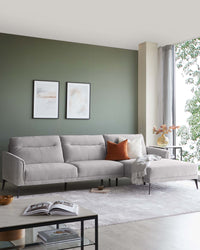 Modern light grey sectional sofa with tufted back cushions and a chaise lounge extension, accented with contrasting orange and white throw pillows and a light grey throw blanket. The black minimalist coffee table with glass top stands in front, beside which lies a hardcover book.