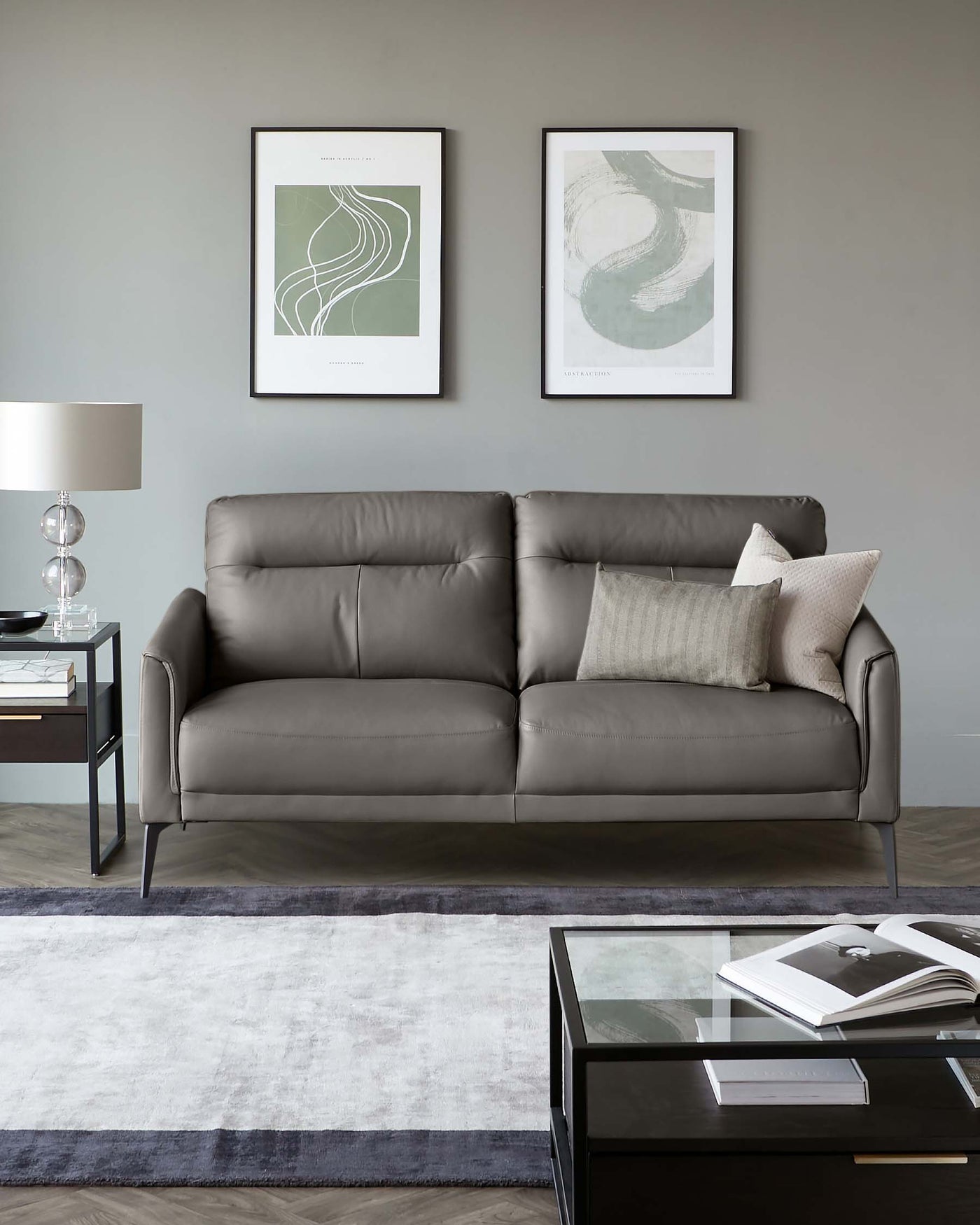 Three-seat leather sofa in a modern design with sleek metal legs, complemented by minimalist side and coffee tables with glass tops and metal frames, accented with a contemporary table lamp, all staged on a two-tone area rug.
