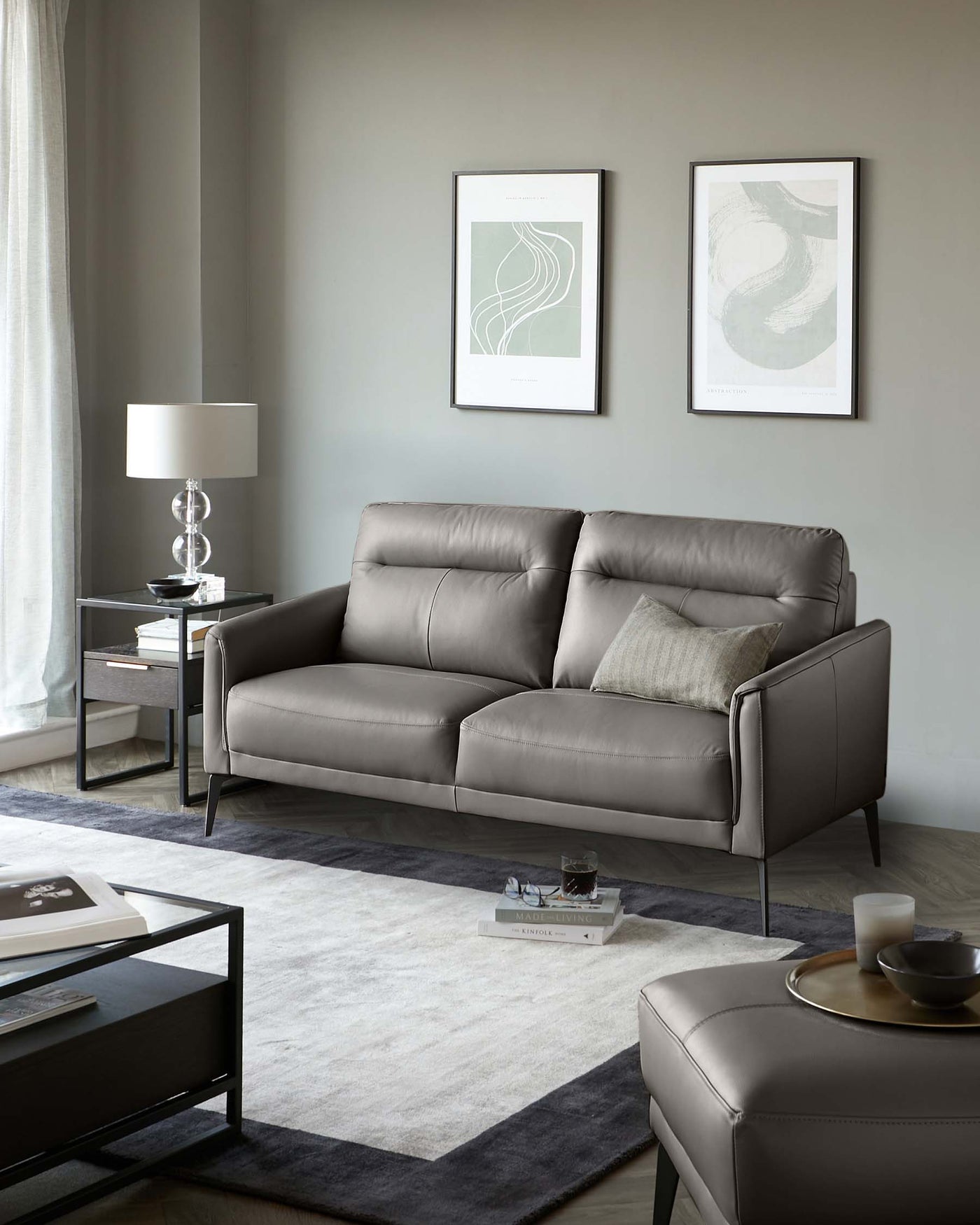 Elegant modern living room furniture featuring a sleek grey leather sofa with clean lines and metal legs, complemented by a matching leather ottoman and minimalist black side tables adorned with contemporary lamps. A modern low-profile coffee table centres the arrangement atop a two-tone grey area rug.