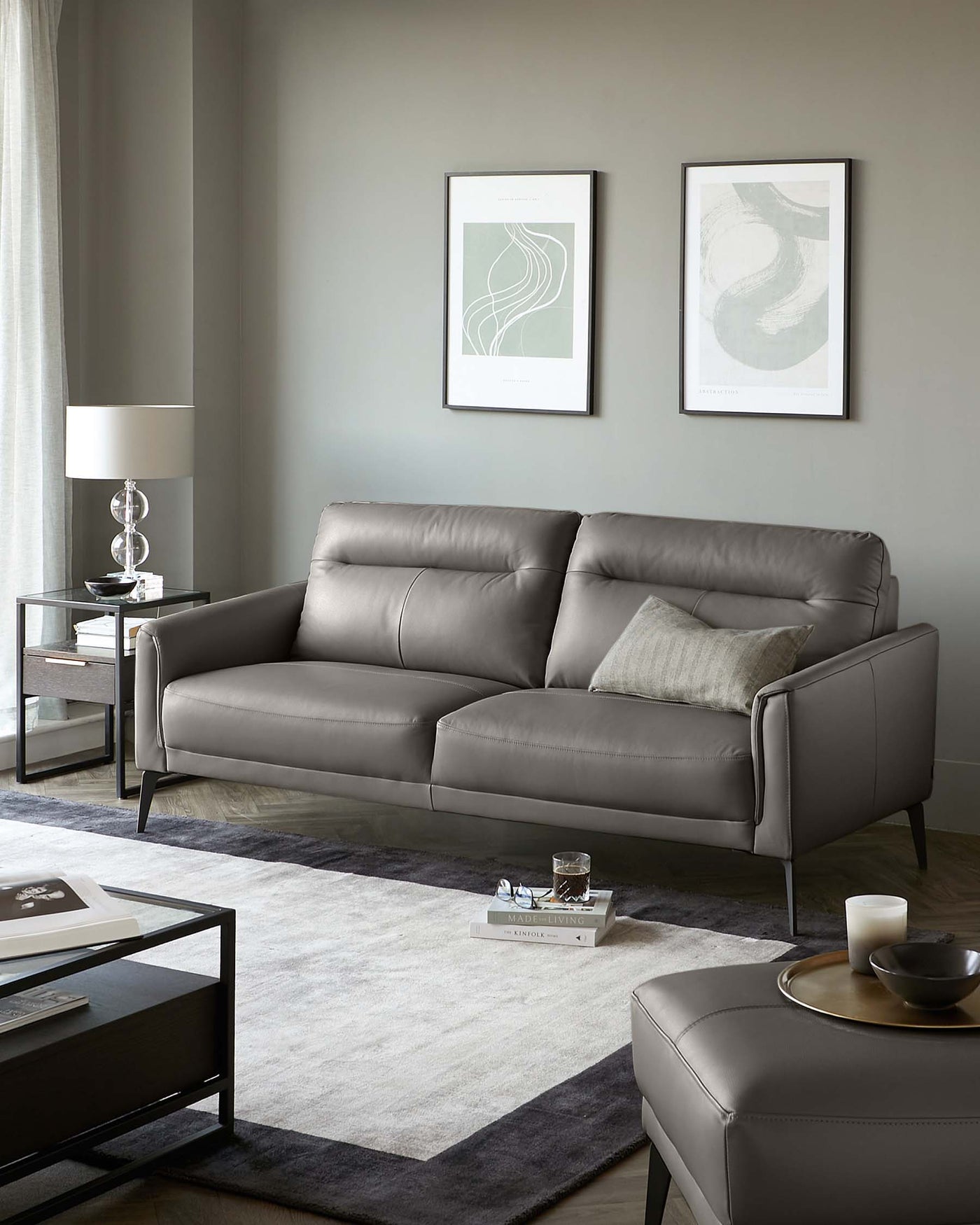 Modern living room setup featuring a sleek grey leather sofa with clean lines and minimalist design, accompanied by a matching leather ottoman acting as a coffee table. A pair of elegant side tables with glass tops and metallic frames flank the sofa, each holding a stylish table lamp. The furniture ensemble rests atop a large two-tone area rug, which adds warmth and texture to the space.