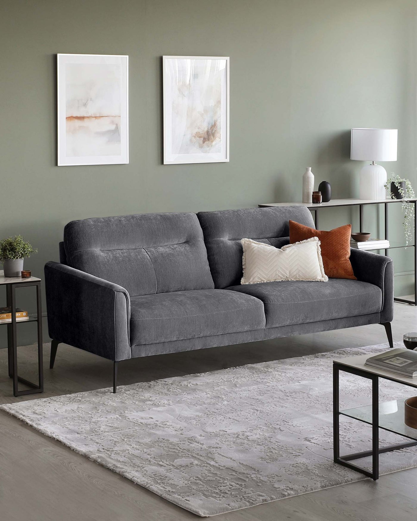 A modern charcoal grey fabric sofa with two seat cushions and black metal legs, complemented by a mix of white and burnt orange throw pillows. In front of the sofa is a light grey patterned area rug. To the side, there is a black metal frame side table with a simple, rectangular design, topped with a small potted plant and books. Another matching table is partially visible, featuring decorative items and books.