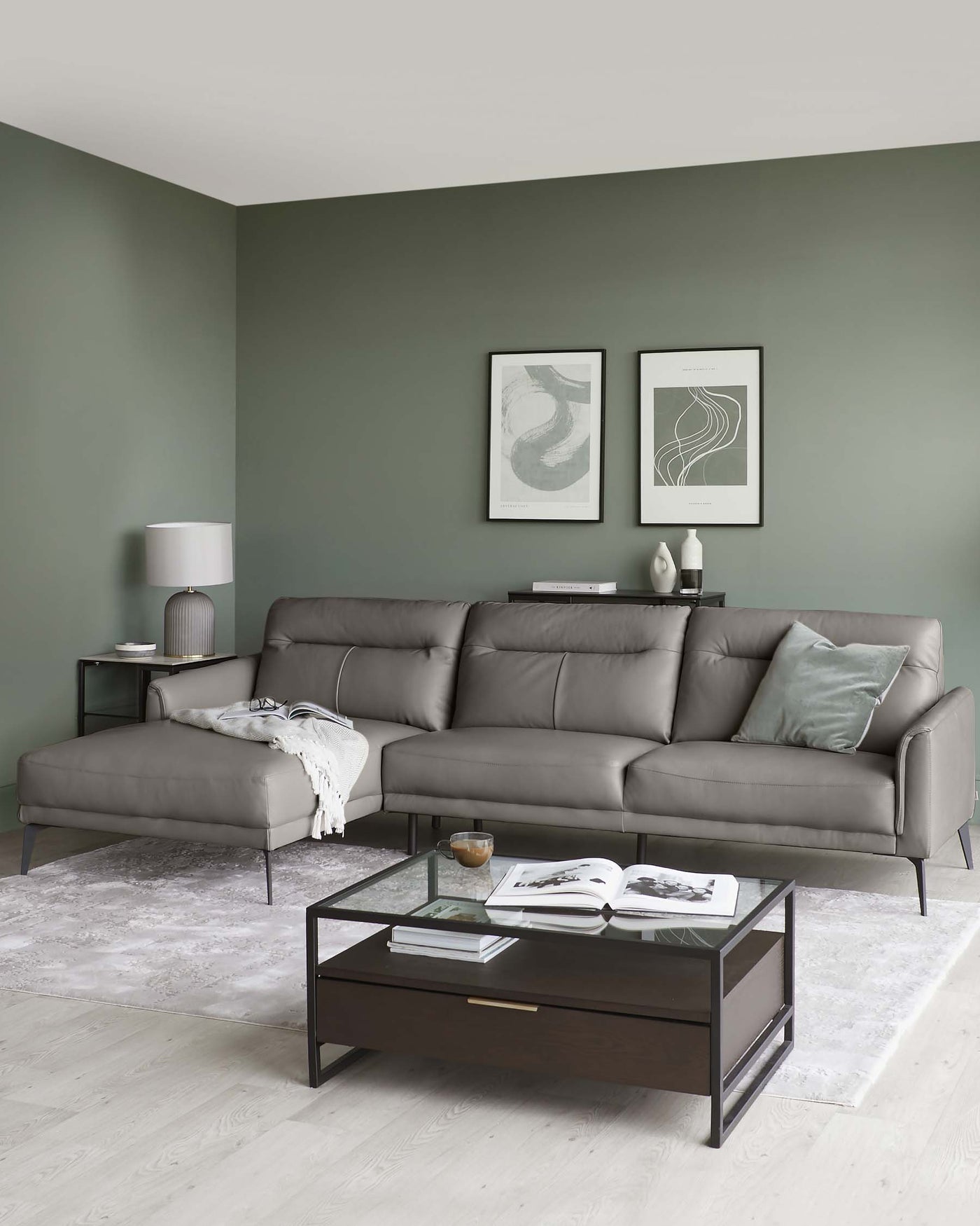 A modern living room featuring a large L-shaped sectional sofa in a muted grey upholstery, flanked by minimalist metal-frame side tables in a matching tone, accompanied by a rectangular dark wood coffee table with a glass top and metal frame. A soft white throw is casually draped over one side of the sofa, and decorative cushions accent the furniture. The room is styled with a subdued colour palette, creating a sophisticated and serene setting.