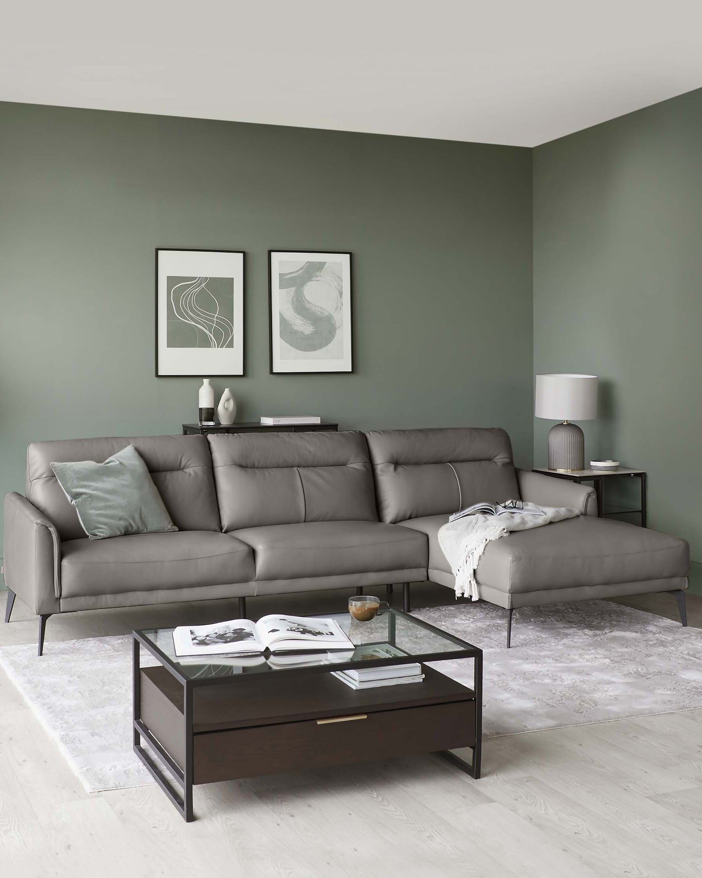 Modern living room featuring a sleek, grey L-shaped sectional sofa with metal legs, a dark wooden coffee table with a clear glass top and an open shelf, paired with a matching wooden side table holding a cylindrical table lamp.