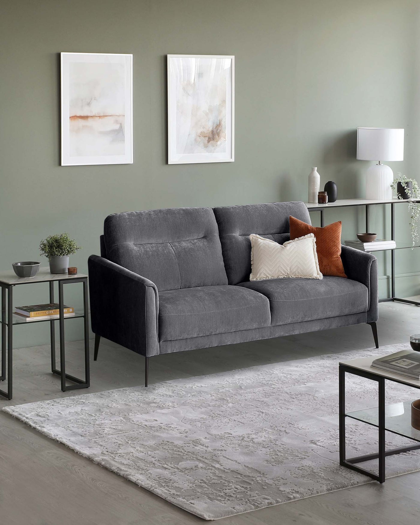 A contemporary, charcoal grey upholstered sofa with a tufted backrest and boxy armrests, featuring neutral-toned accent pillows. Positioned on a textured ivory area rug, the setting includes two minimalist black metal end tables with glass tops and a matching coffee table with a clear glass inset and a lower shelf. An understated off-white table lamp with a cylindrical shade adorns one end table, complementing the modern aesthetic of the room.