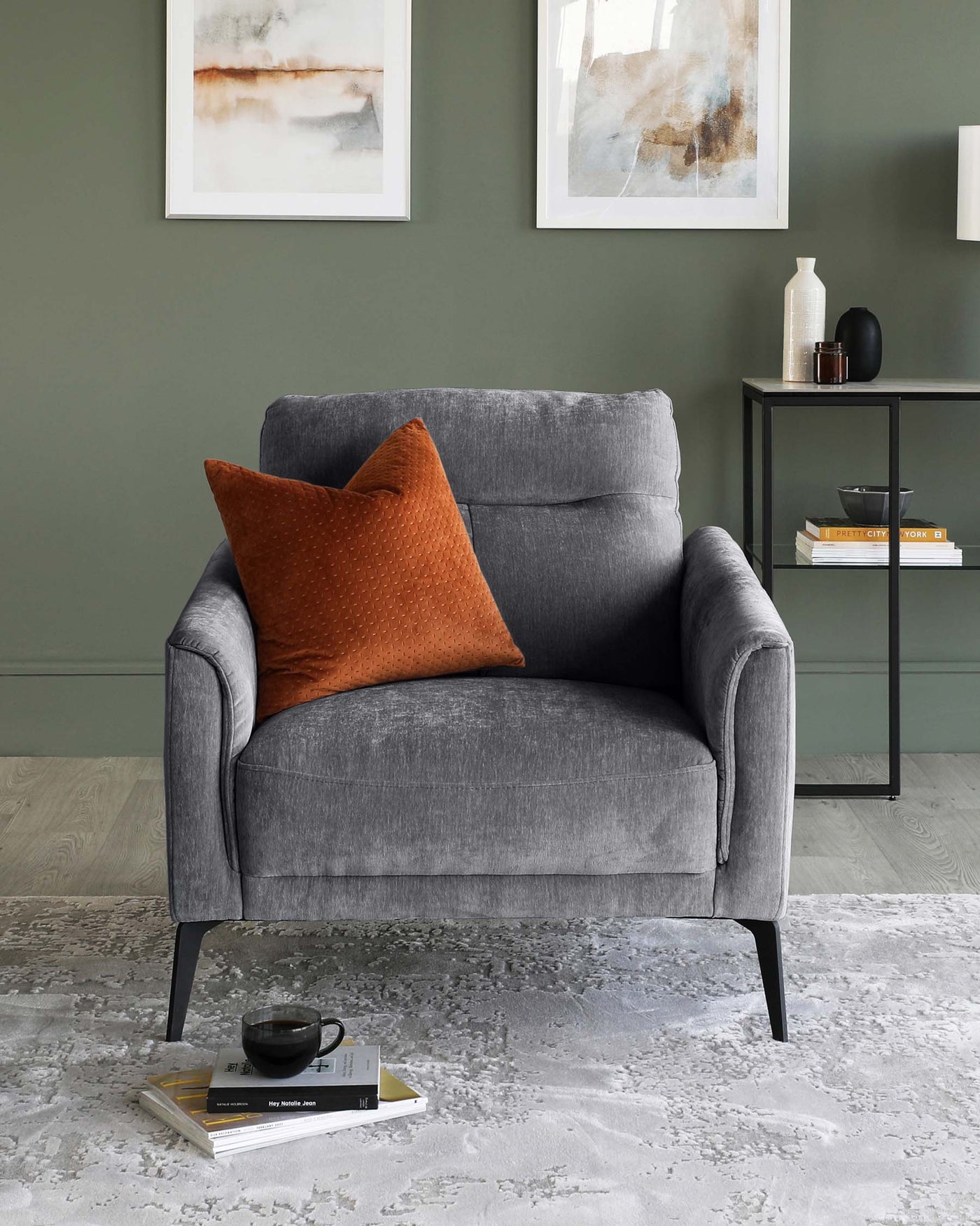 A modern grey fabric armchair with black metal legs, featuring a curved backrest and a plush seat, complemented by a single orange textured throw pillow. In the setting, there is also a minimalist matte black metal side table with a rectangular surface and a lower shelf holding books and decorative items.