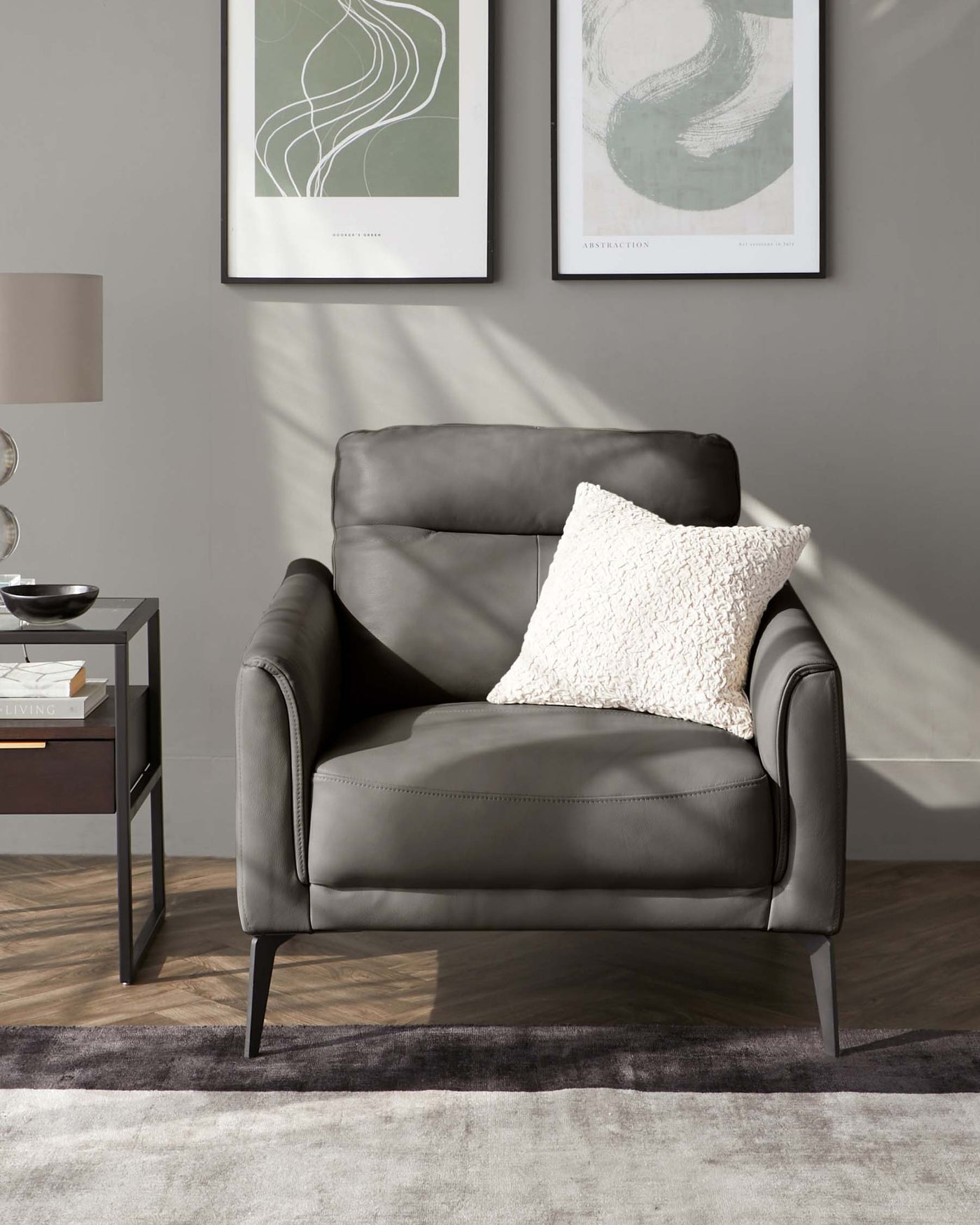 Modern charcoal leather armchair with clean lines and metal legs, complemented by a white textured throw pillow, set against a grey wall with framed abstract art and beside a minimalist black end table with decorative items. There's a grey floor lamp and a plush area rug completing the stylish scene.