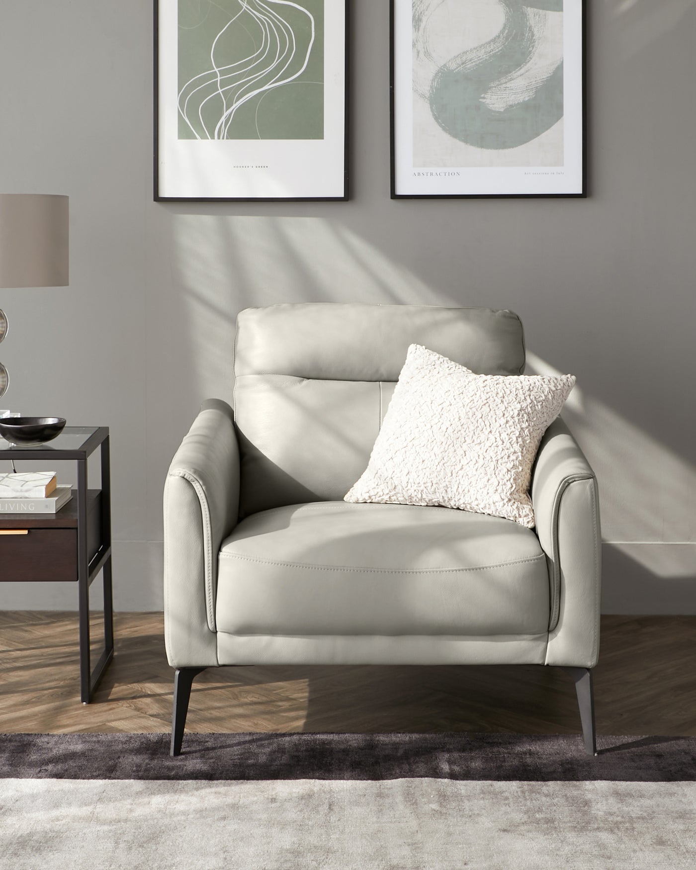 Modern light grey leather armchair with sleek lines and black metal legs, accompanied by a textured white throw pillow. Beside it, a contemporary black side table with geometric lines and a matte surface, topped with a bowl decor. The setting includes abstract wall art, a floor lamp, and a plush area rug over herringbone wood flooring.