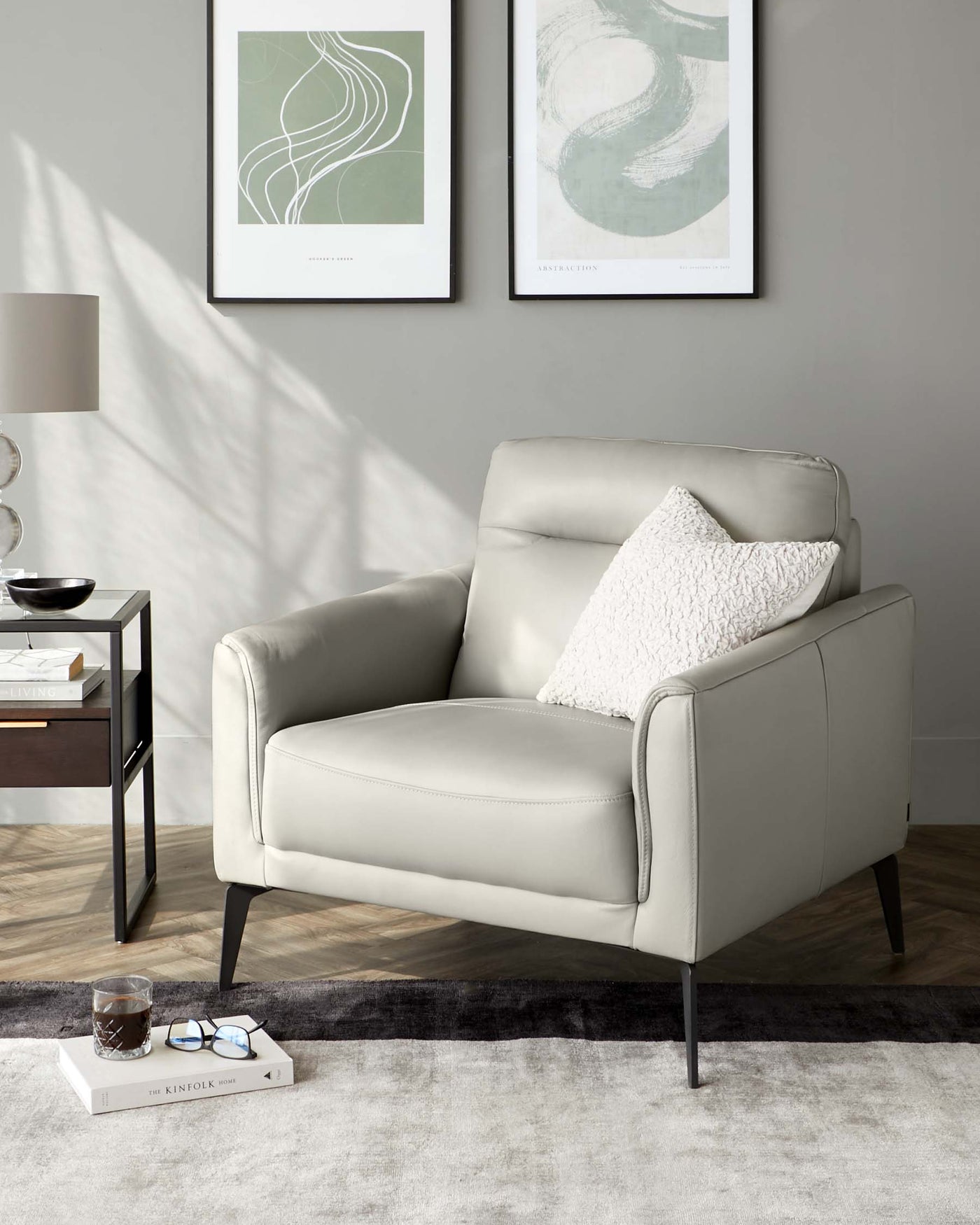 Elegant light grey leather armchair with a modern silhouette, featuring clean lines, a cushioned seat, a comfortable backrest, and a fluffy white throw pillow. The chair has slender black metal legs and is placed beside a contemporary style side table with a round black top and a lower wooden shelf, accessorized with decorative items. The setup is on a plush dual-tone grey area rug over a hardwood floor, with abstract wall art in the backdrop.