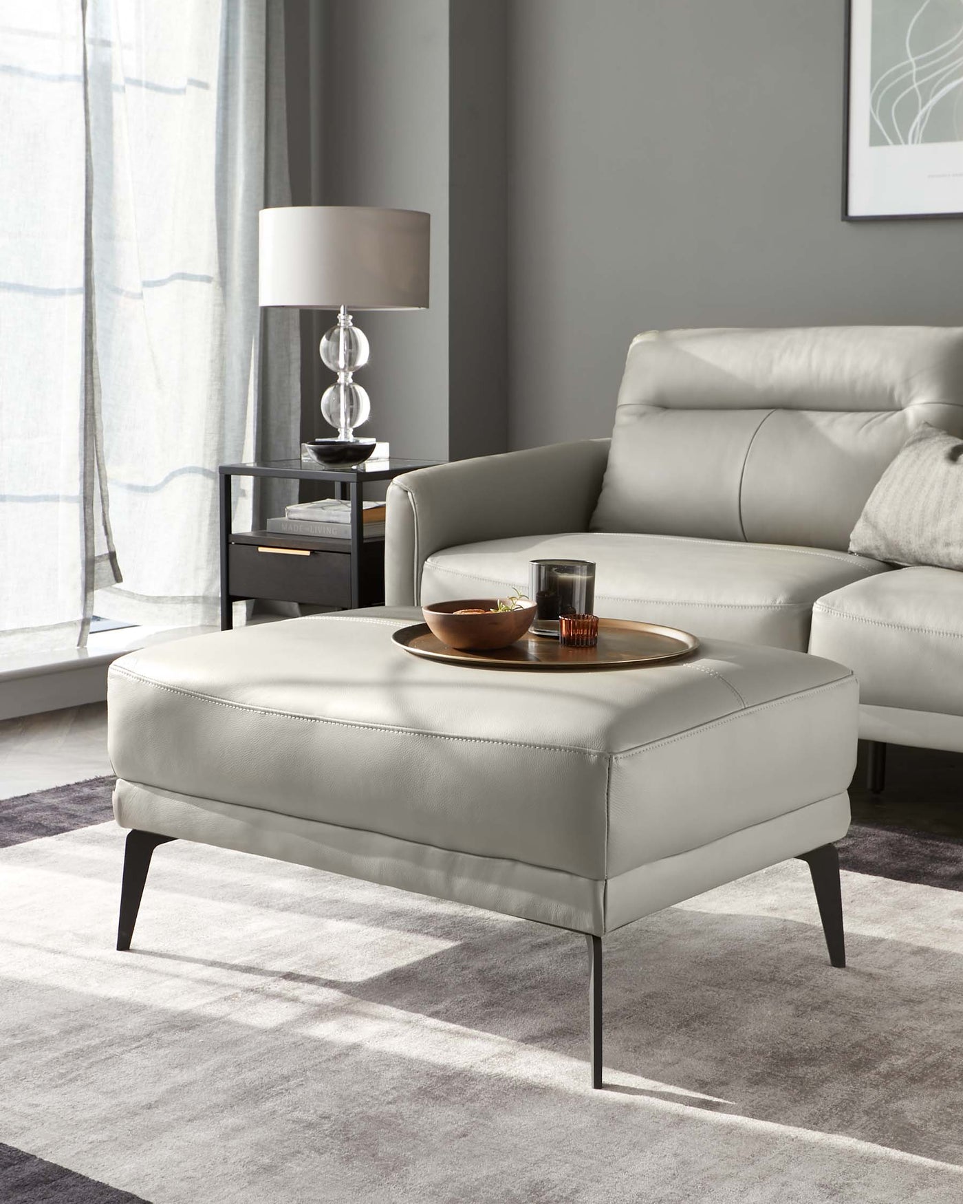 Contemporary light grey leather armchair and matching ottoman with slender dark metal legs, paired with a minimalist black metal side table featuring a sleek table lamp, set on a soft grey area rug.