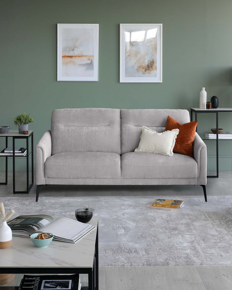 A modern living room scene featuring a minimalist, elegant three-seater sofa with a neutral grey fabric upholstery and sleek black metal legs. Accompanying the sofa is a set of two matching side tables in black with square tops and a coordinating rectangular coffee table, all displaying a selection of decorative items. The room is adorned with a light grey area rug and a pair of abstract wall paintings, enhancing the contemporary aesthetic.