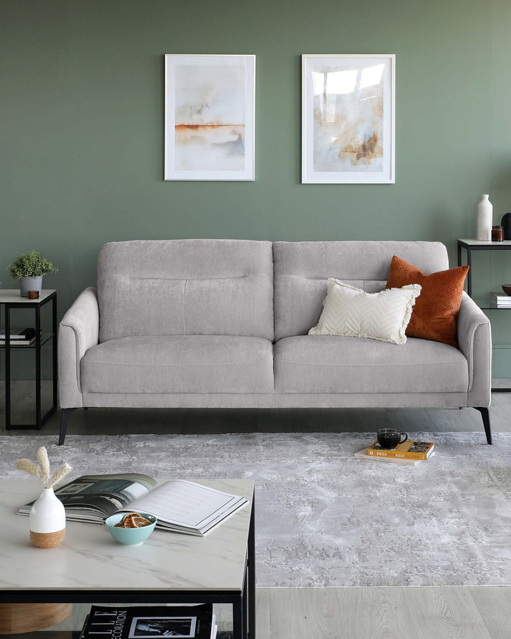 A modern, minimalist living room featuring a light grey fabric three-seater sofa with a tufted back, slim armrests, and dark slender legs. Accompanying the sofa are two square side tables, one black metal with a glass top and the other matching with a lower shelf, displaying small decorative items and books. In front of the sofa is a rectangular marble-topped coffee table with a black metal frame, holding a book, vase, and small bowl. The colour palette is neutral with monochrome and earth-toned accents.