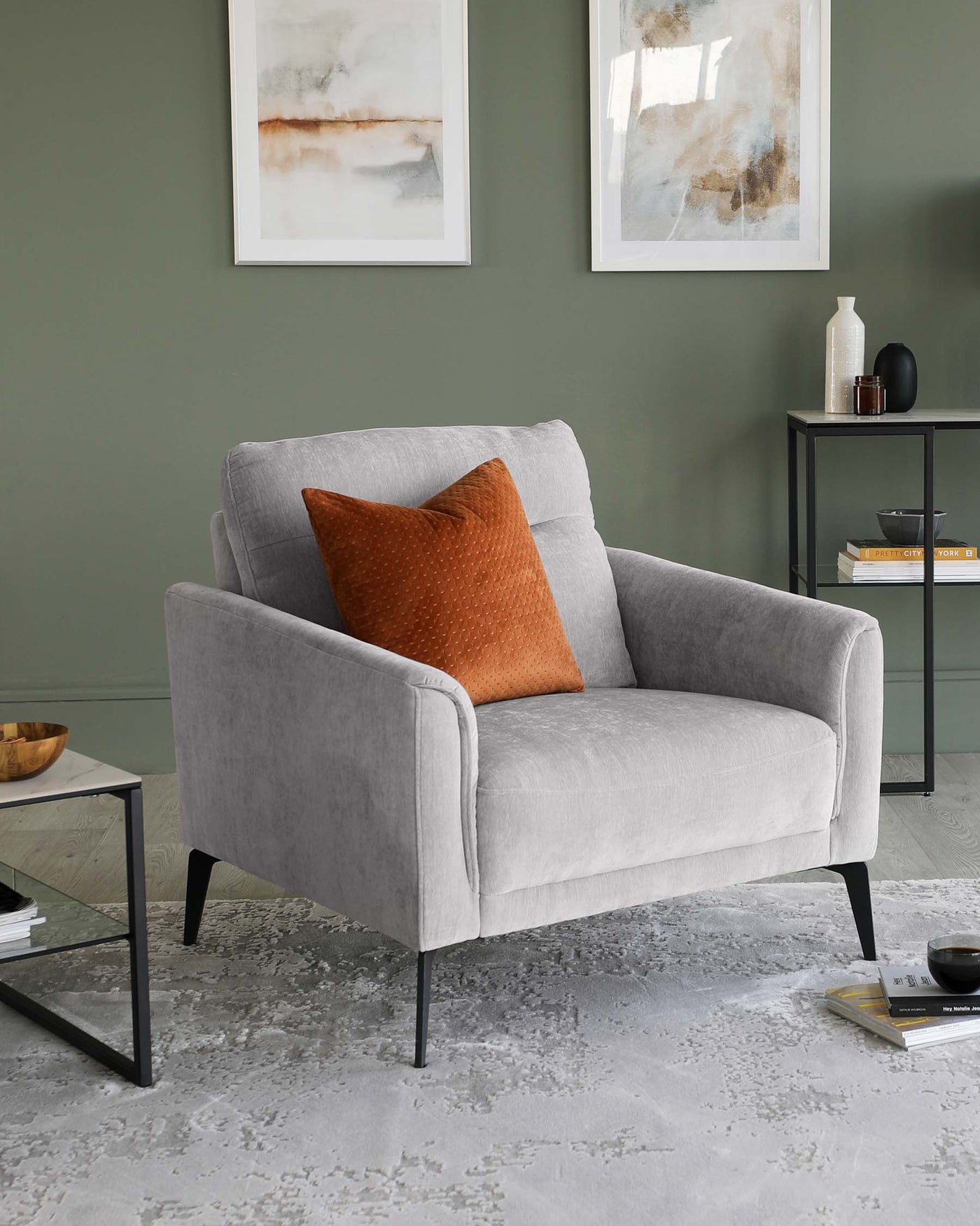 Elegant modern armchair with light grey upholstery and black legs, accompanied by a burnt orange decorative pillow. Adjacent is a minimalist black metal side table with a simple vase, a black decorative object, and a stack of books. The setting is complemented by abstract wall art and a textured grey area rug.