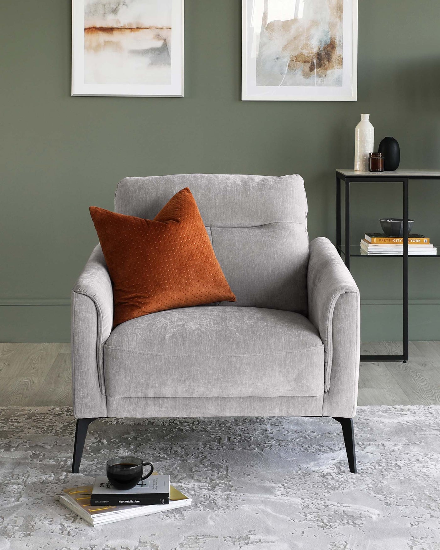 Modern, minimalist-style armchair in light grey fabric with textured rust-coloured accent pillow. The chair has a simple, clean design featuring a high backrest and curved armrests, resting on slender, angled black-metal legs. Beside it is a sleek black-metal side table with a rectangular top, displaying decor items and books. The setup is on a textured light grey area rug over a hardwood floor.