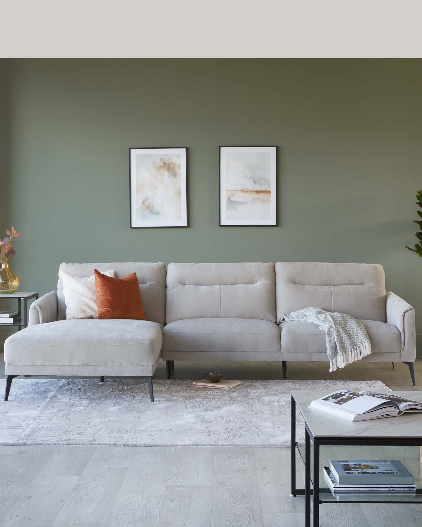 Modern light grey fabric sectional sofa with clean lines and metal legs, accompanied by a matching chaise lounge, positioned on a textured off-white area rug. A sleek black metal coffee table with a glass top and magazines on the lower shelf is placed in front. The ensemble is framed with minimalist wall art above the sofa and a small indoor plant to the left, creating an elegant living space.