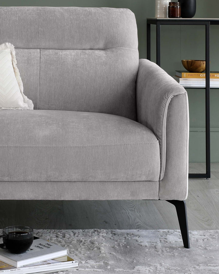 A modern light grey fabric sofa with clean lines, featuring cushioned armrests and a plush backrest. The sofa is styled with a textured white throw pillow and stands on elegant tapered black legs. In the background, a sleek black metal and glass shelving unit with various decorative items and books is partially visible.