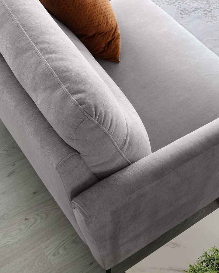 Contemporary grey fabric sofa with plush cushioning and a single brown decorative pillow, featuring streamlined armrests and a soft, smooth upholstery finish, against a backdrop with a wooden floor and a partial view of a grey rug.