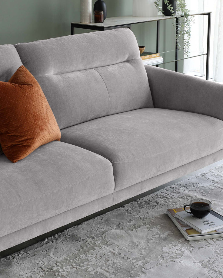 A contemporary grey fabric sofa with clean lines and a plush finish, featuring a single burnt orange textured throw pillow. The sofa sits on a subtle metal base, positioned on a textured grey and white area rug, with a sleek black metal and glass shelving unit in the background housing decorative items and books. A coffee cup and saucer rest atop a hardcover book on the rug near the sofa.