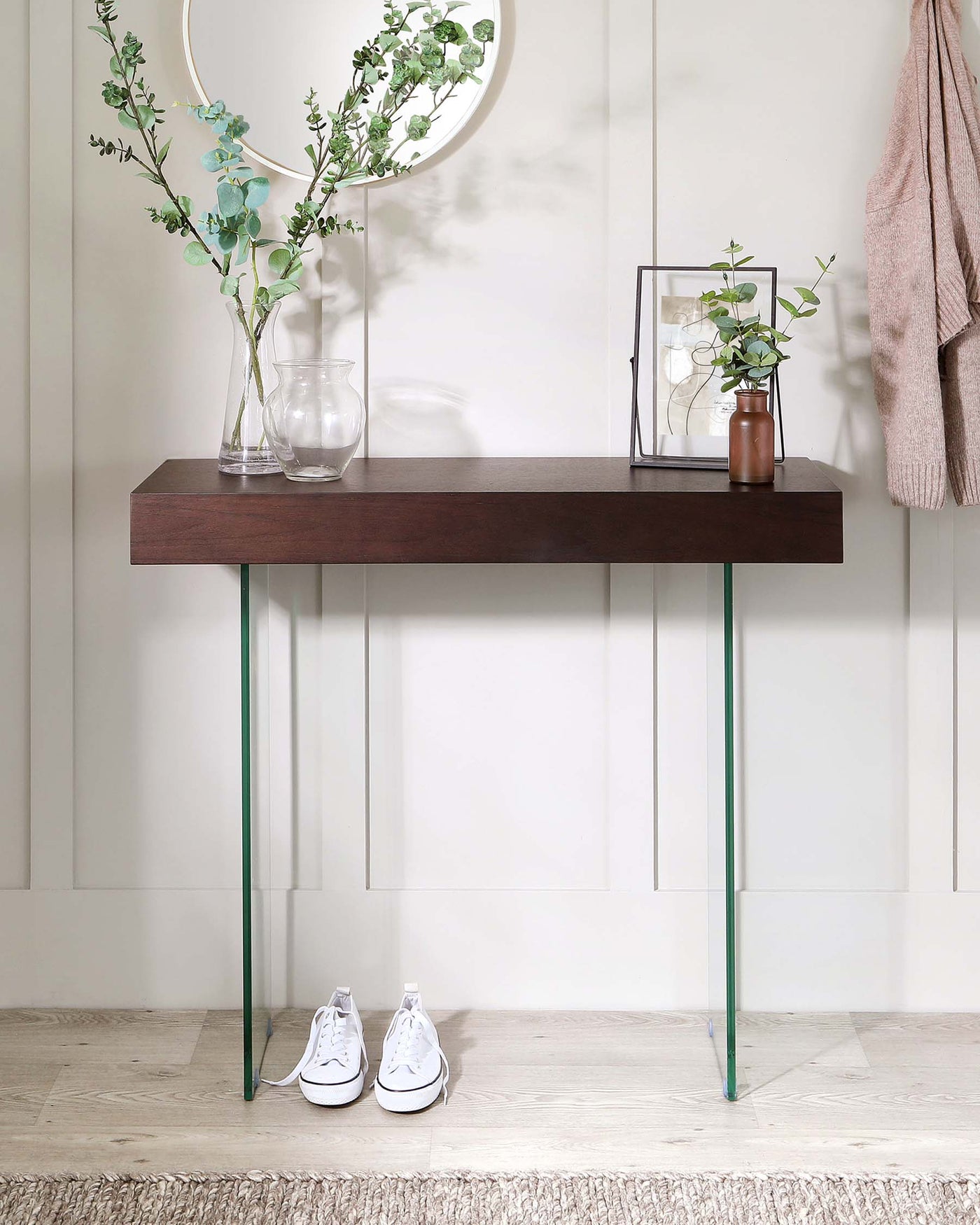 Modern minimalist dark wooden console table with clear glass legs. The table is adorned with decorative items including a tall vase with greenery, a spherical mirror above it, a glass candle holder, and a small plant in a brown vase. A pair of white sneakers is placed neatly below the table, on a light wooden floor adjacent to a textured rug.