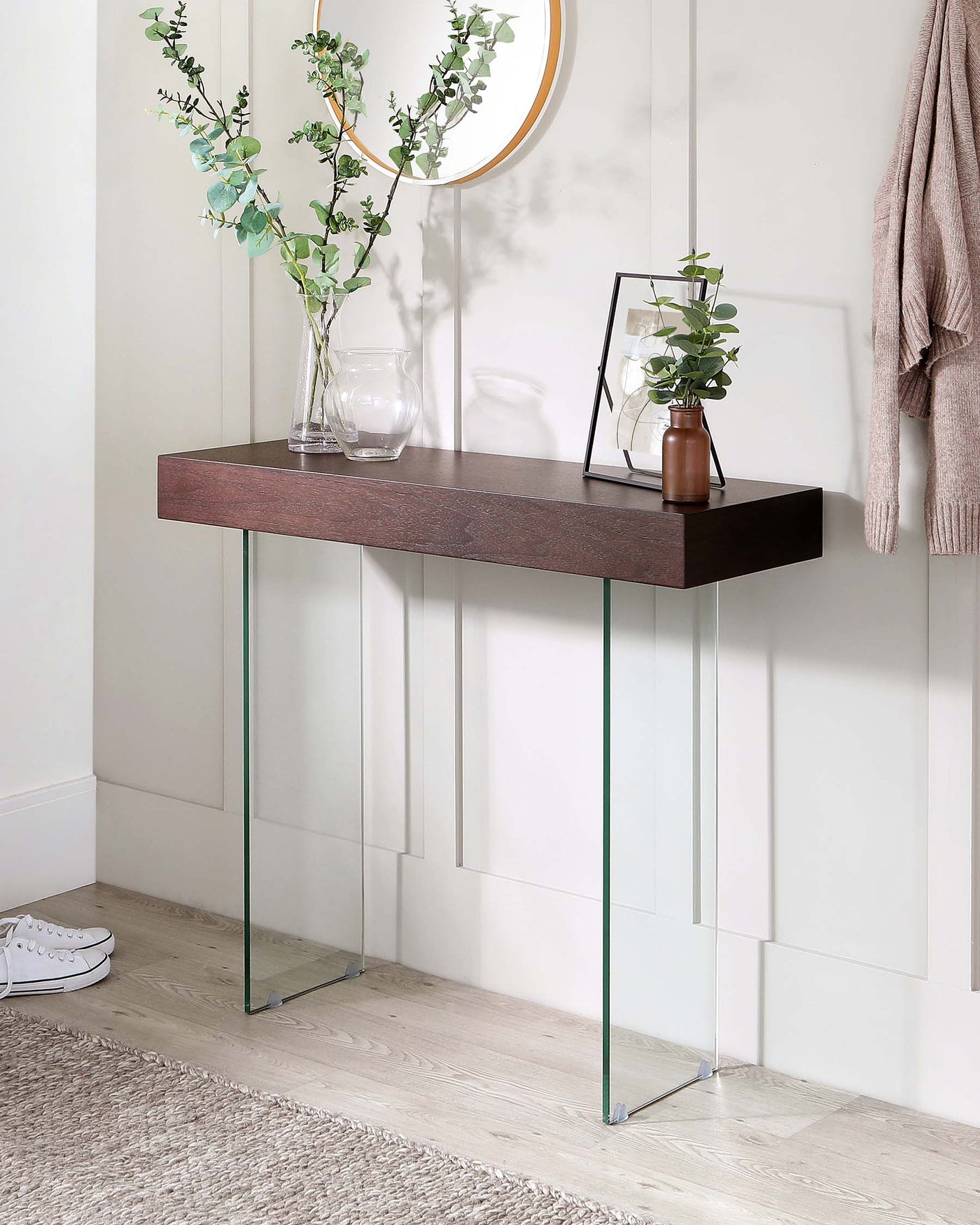 A modern minimalist console table with a rich dark brown wooden tabletop and sleek, clear glass legs. The tabletop is styled with decorative items including a vase containing greenery, a glass candle holder, a picture frame, and a small potted plant. The console sits against a light wall above a light wood floor, complemented by a round mirror and a light beige throw hanging beside it.