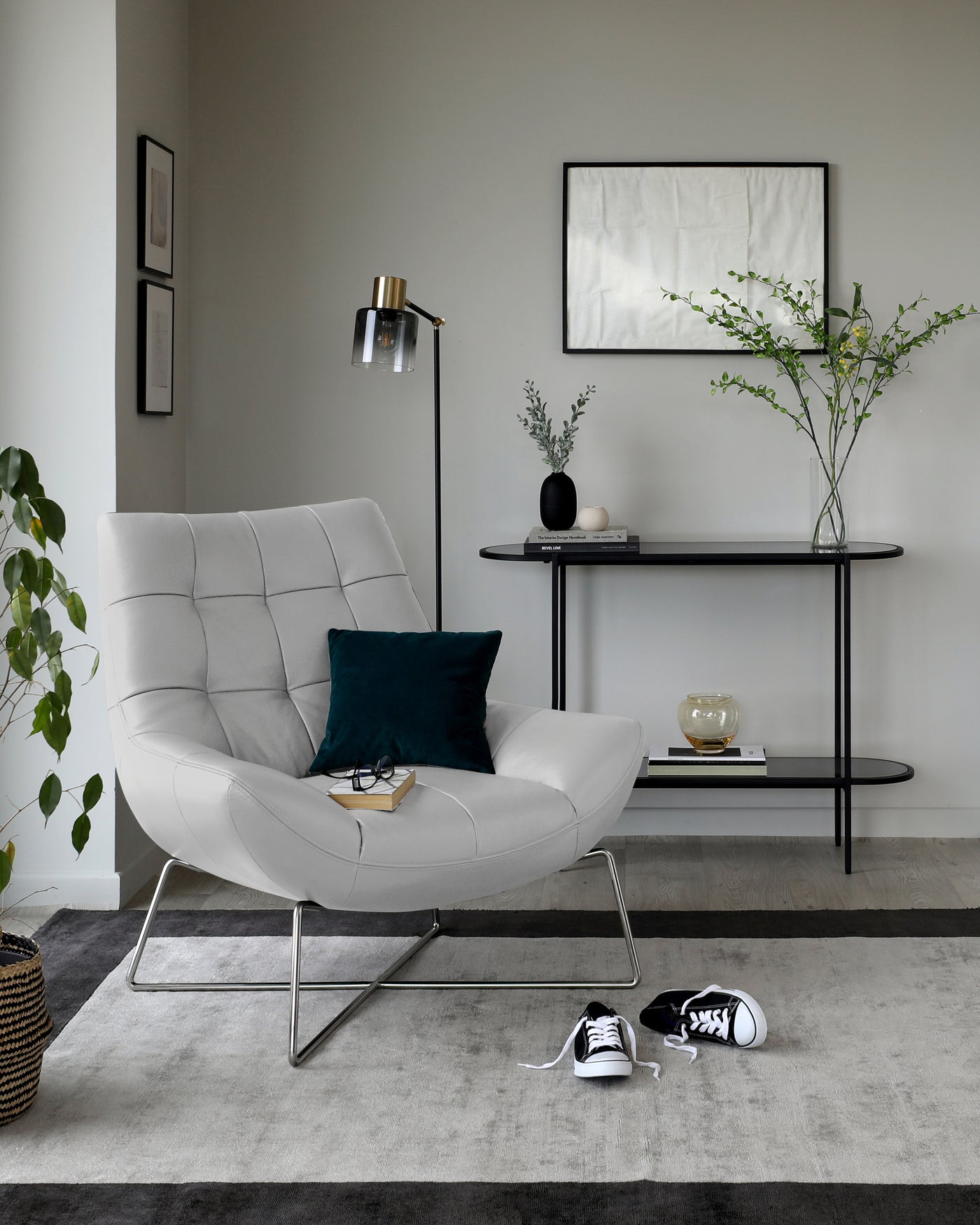 Modern light grey upholstered armchair with a unique curved design and slim silver metal legs. Behind it is a sleek, round-edged rectangular black console table with thin metal legs, adorned with minimalistic decorative items. Both pieces complement a contemporary interior aesthetic.
