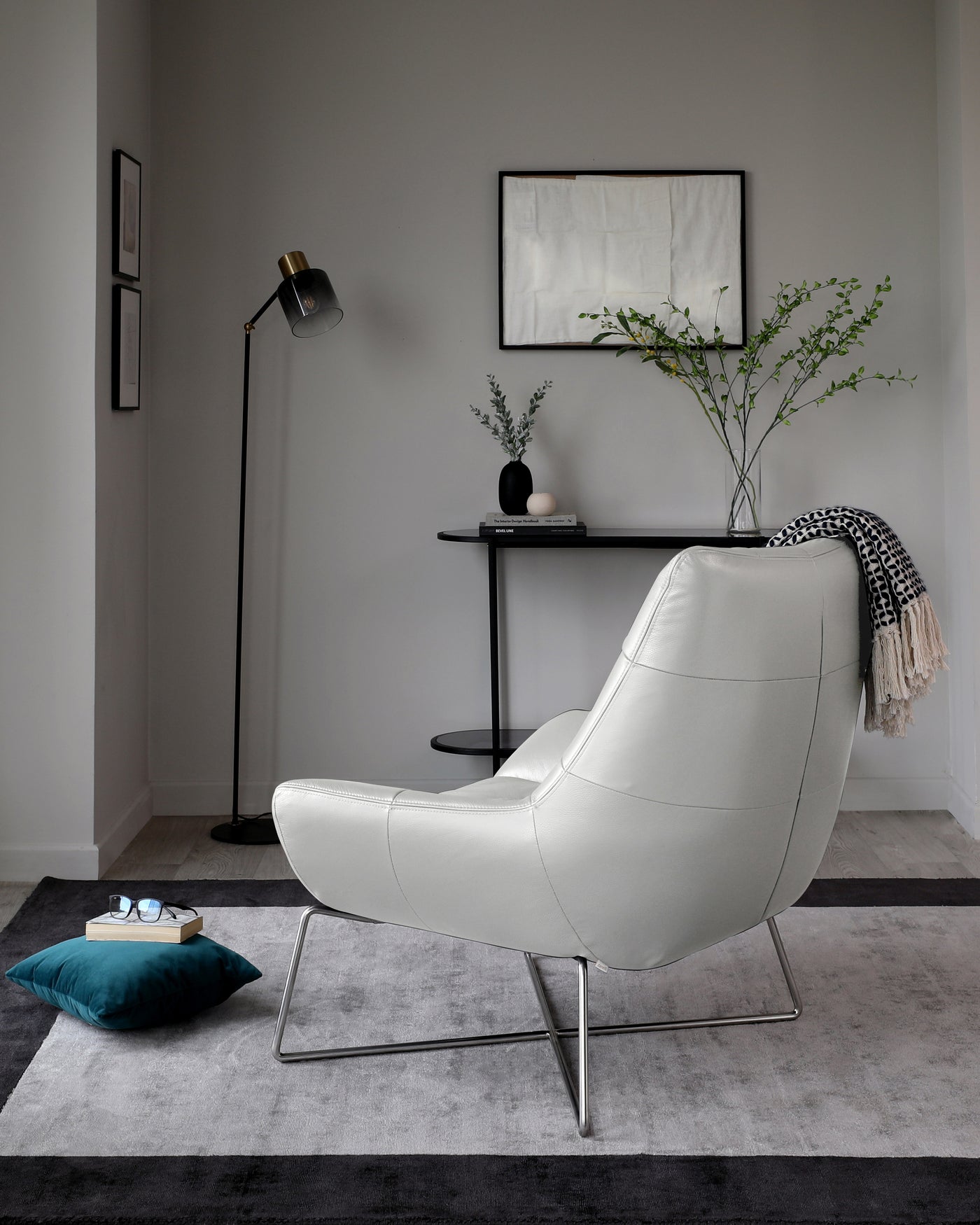 Modern light grey leather lounge chair with a high back and a chrome base, paired with a round black side table with two tiers, displaying decor and greenery. On the side, a tall black floor lamp with a minimalist design.