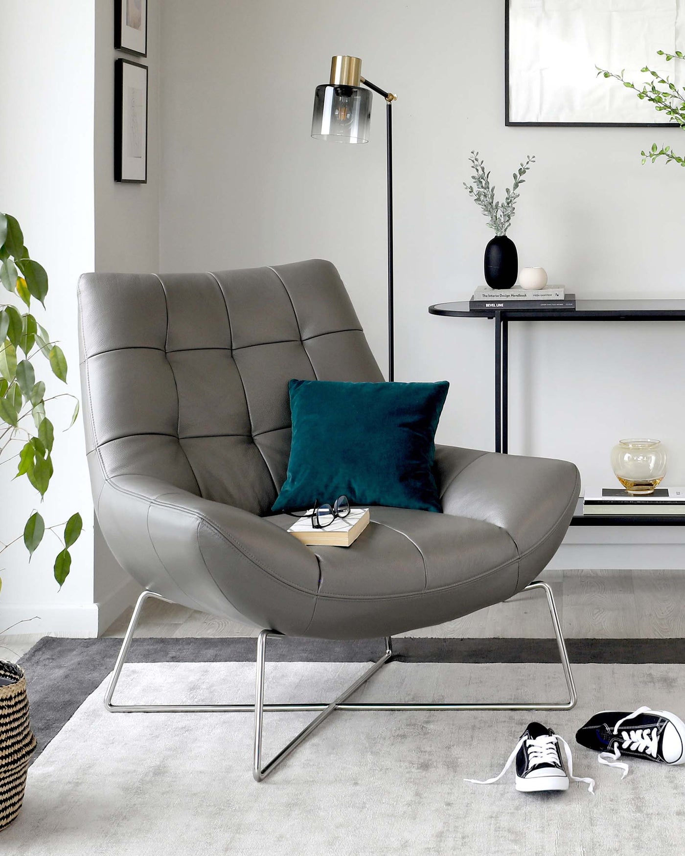 Modern grey leather upholstered lounge chair with tufted backrest and cushioned seat, supported by a sleek, chrome-finished metal frame with cantilever base. Beside it is a stylish black floor lamp with a gold accent and a glass shade, alongside a minimalist black console table holding decorative items.