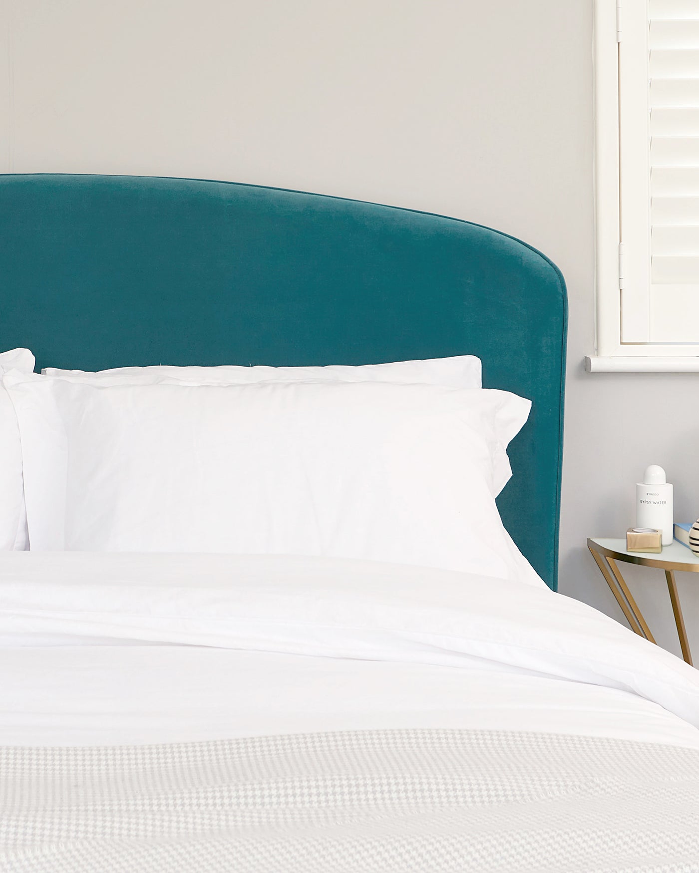 Elegant teal velvet upholstered headboard with a smooth curved top, paired with a clean white bedding set on a minimalist bed frame, adjacent to a simple wooden side table with a round top and triangular base.