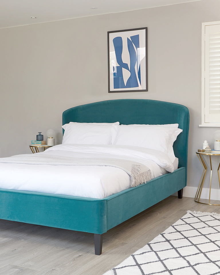 Elegant teal upholstered bed with a curvilinear headboard and dark wooden legs, accompanied by a gold and glass side table. The bed is dressed in white linens and a light grey throw. A geometric patterned rug lies on the light wooden floor.