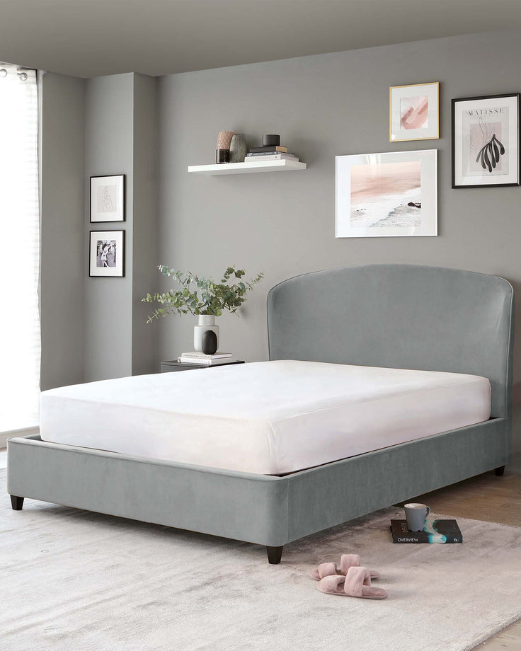 Elegant upholstered bed frame in a soft grey fabric with a high, gently curved headboard and low-profile footboard, supported by dark wooden legs, displayed in a well-appointed bedroom setting.