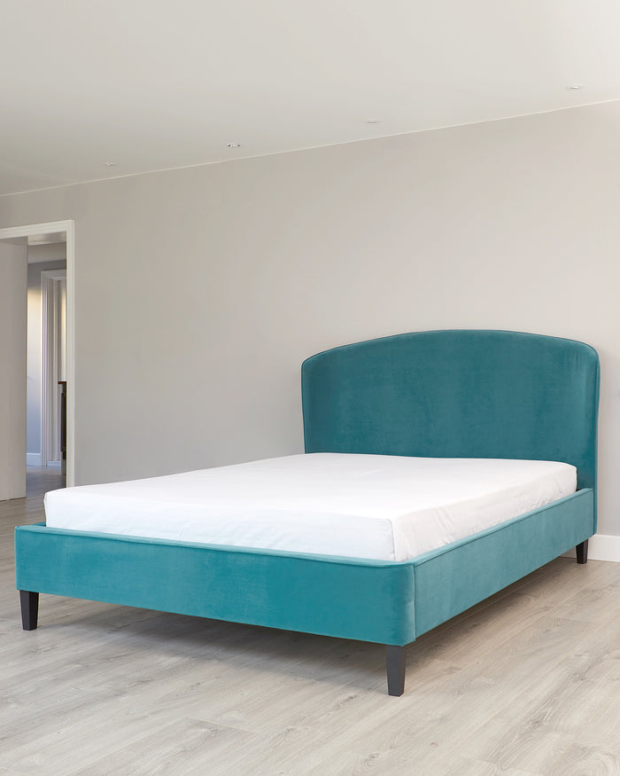 Elegant teal upholstered bed frame with a curved headboard and dark wooden legs, set against a light wooden floor and neutral background.