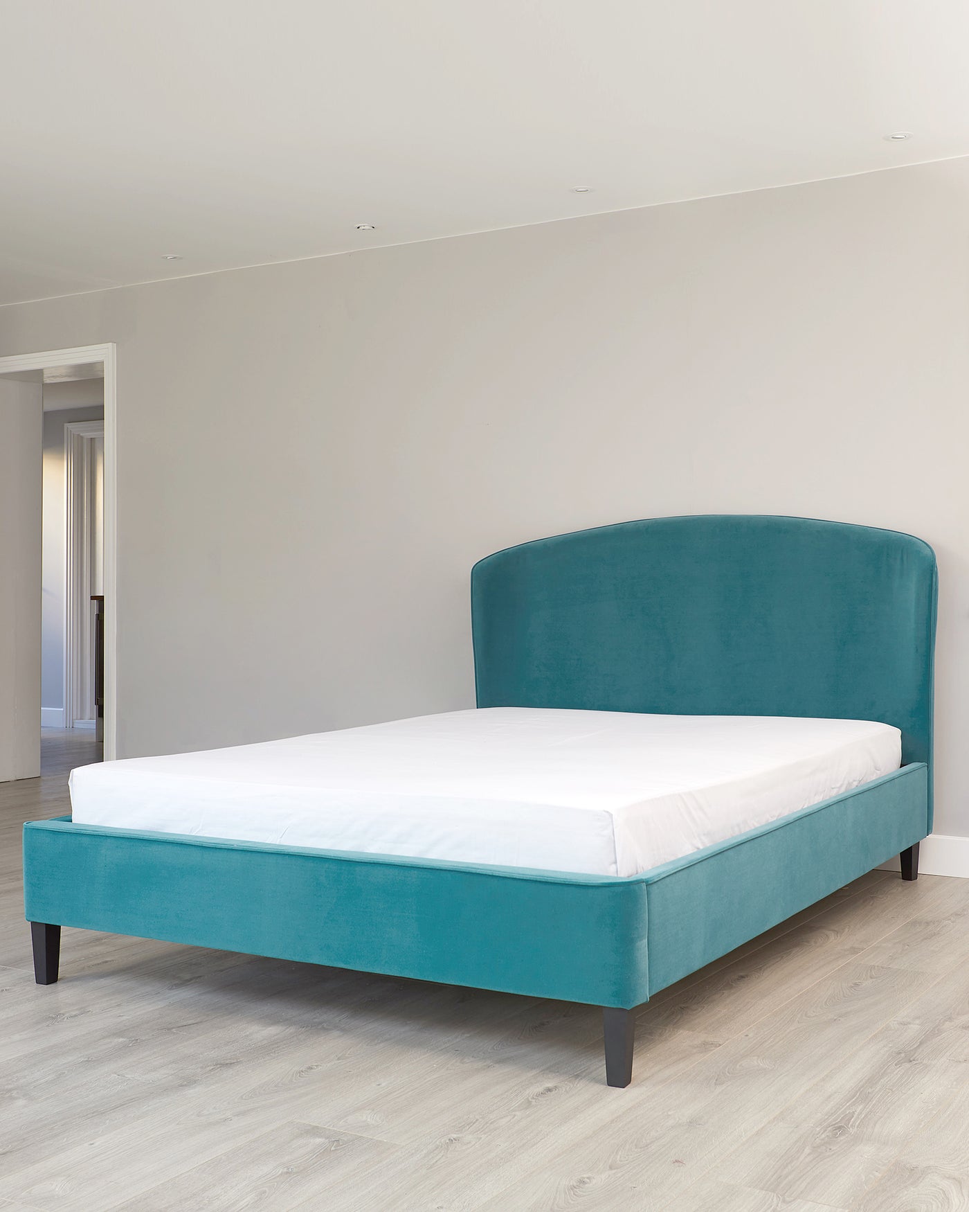 A modern teal upholstered platform bed with a curved headboard and dark wooden legs, showcasing a simplistic design ideal for contemporary bedrooms. The bed is dressed with a plain white mattress, emphasizing clean lines and a minimalist aesthetic.