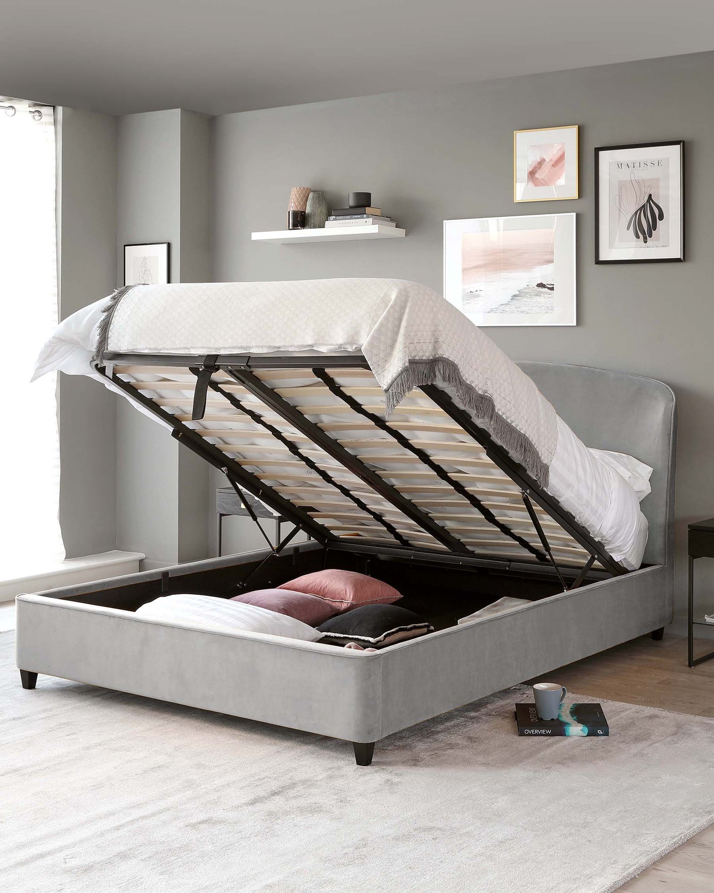 Gray upholstered storage bed with lifted mattress platform revealing under-bed storage space, set in a room with neutral decor and wall art.