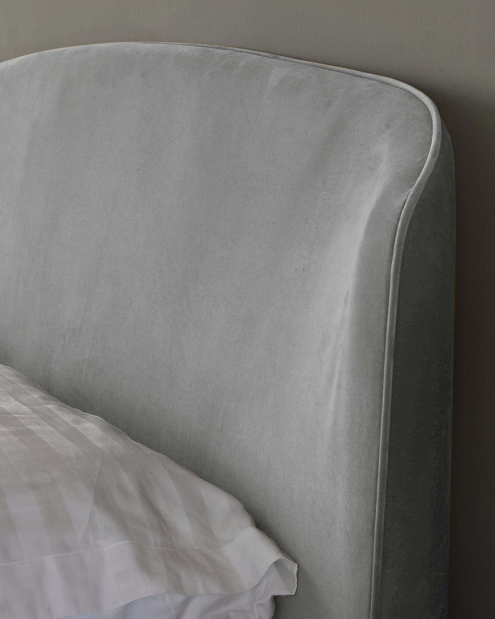 Close-up of a modern styled, velvet upholstered bed headboard in a soft grey colour with a subtle sheen, paired with a glimpse of crisp white bed linen.