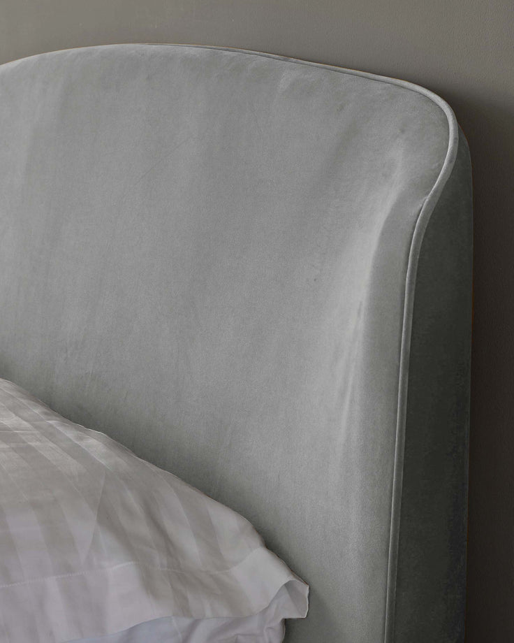 Close-up of a modern styled, velvet upholstered bed headboard in a soft grey colour with a subtle sheen, paired with a glimpse of crisp white bed linen.