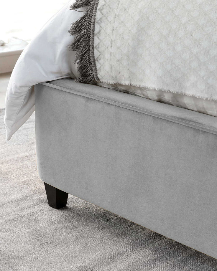 An elegant light grey upholstered bed frame with a streamlined silhouette and minimalist black block feet, partially covered by a white comforter with a decorative fringe throw. The bed is placed on a soft grey carpet, suggesting a modern and serene bedroom setting.