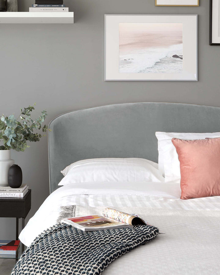 A modern bedroom featuring a king-sized bed with a plush, curved light grey fabric headboard and crisp white bedding. Beside the bed is a small, black contemporary nightstand with a round vase and stacked books. A cosy blue and white knitted throw is casually draped across the bed.