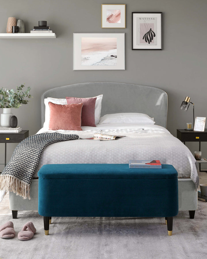 Elegant contemporary bedroom furniture featuring a plush upholstered queen-sized bed with a curved headboard design in a soft grey fabric. Accompanying the bed is a striking teal blue velvet bench with a sleek, rectangular shape and black legs with gold feet caps, providing a pop of rich colour and practical seating at the foot of the bed.