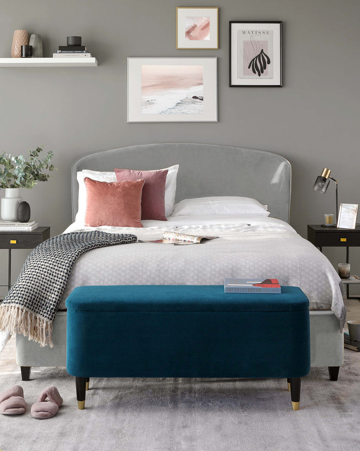 Elegant contemporary bedroom furniture featuring a plush upholstered queen-sized bed with a curved headboard design in a soft grey fabric. Accompanying the bed is a striking teal blue velvet bench with a sleek, rectangular shape and black legs with gold feet caps, providing a pop of rich colour and practical seating at the foot of the bed.