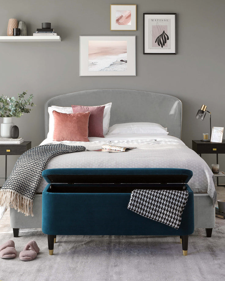 Elegant bedroom setup featuring a curved, upholstered headboard in a soft grey fabric, complementing the clean white bedding. In the foreground, there is a plush, deep teal blue velvet bench with storage capacity at the foot of the bed, accented with brass-capped legs. The room also includes a minimalistic black side table and wall-mounted shelves displaying tasteful decor and artwork.