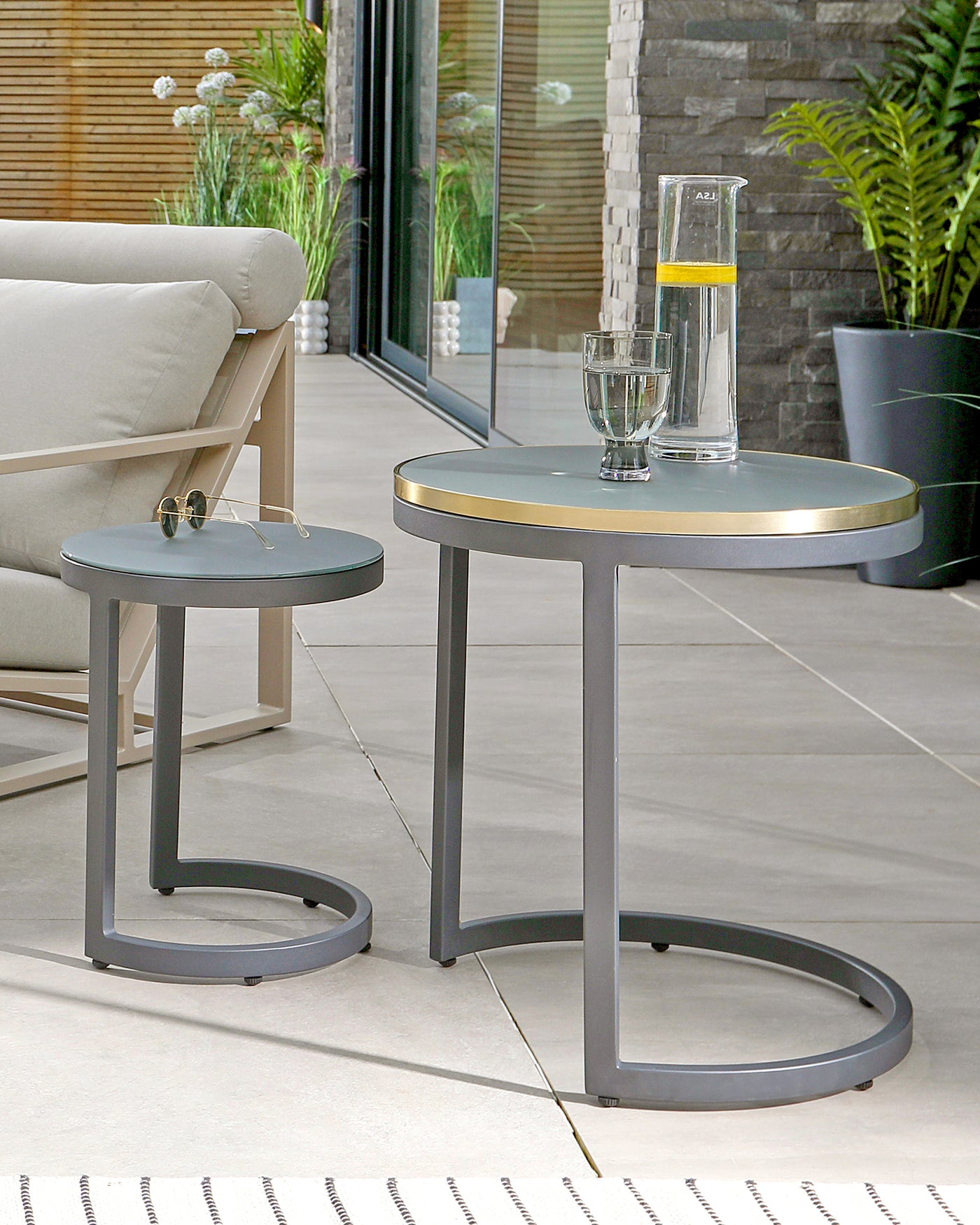 Two modern round side tables with sleek metal frames and a matte finish. The smaller table features a dark grey top, while the larger one has a taupe top with a metallic gold rim. Both have a distinctive C-shaped base that allows for easy integration with various seating arrangements.