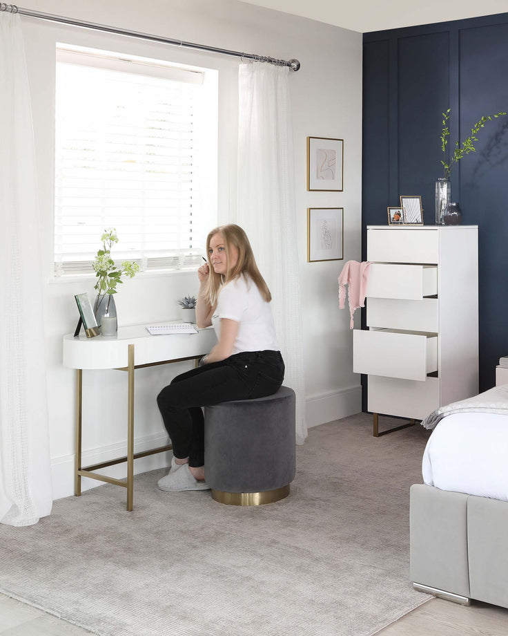 A modern bedroom with a sleek white floating desk paired with a circular grey velvet stool featuring a gold base. The room includes a tall white dresser with clean lines and simple handles, and a light grey upholstered bed with a minimalistic headboard. The furniture is complemented by a pale grey area rug and accented with understated wall art and plants.