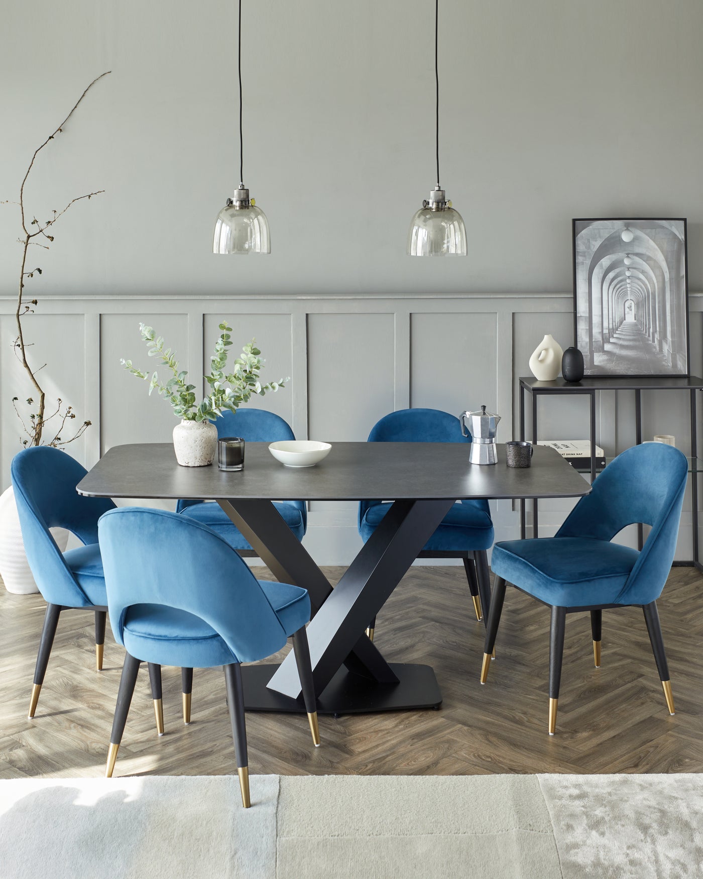 Modern dining room featuring an elegant rectangular black dining table with a unique X-shaped base design. Accompanied by six plush blue velvet dining chairs with black legs tipped with brass accents. The simple yet sophisticated set is complemented by two clear glass pendant lights above the table.