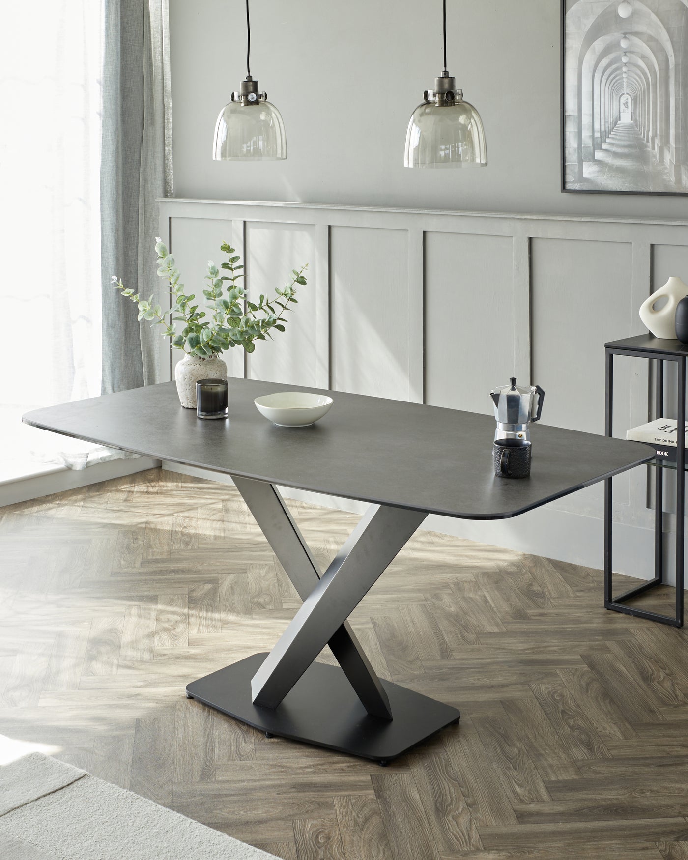 Modern dining table featuring a dark stone or concrete-style tabletop with bevelled edges and a unique, twisted X-shaped metallic base in a matte black finish. The table displays a minimalist aesthetic suitable for contemporary interior design spaces.