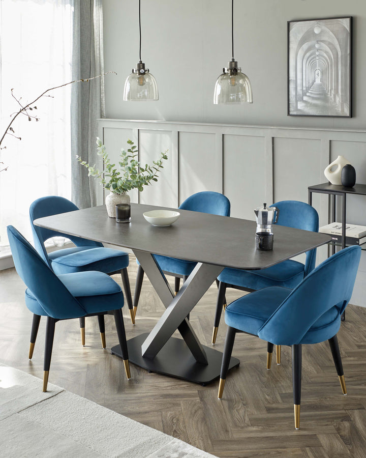 Modern dining room furniture, featuring a rectangular dark grey table with a unique x-shaped metallic base, surrounded by six plush velvet dining chairs in a rich blue tone with black legs tipped in gold. A black sideboard complements the set, along with muted grey walls, herringbone wooden flooring, and ambient pendant lighting.