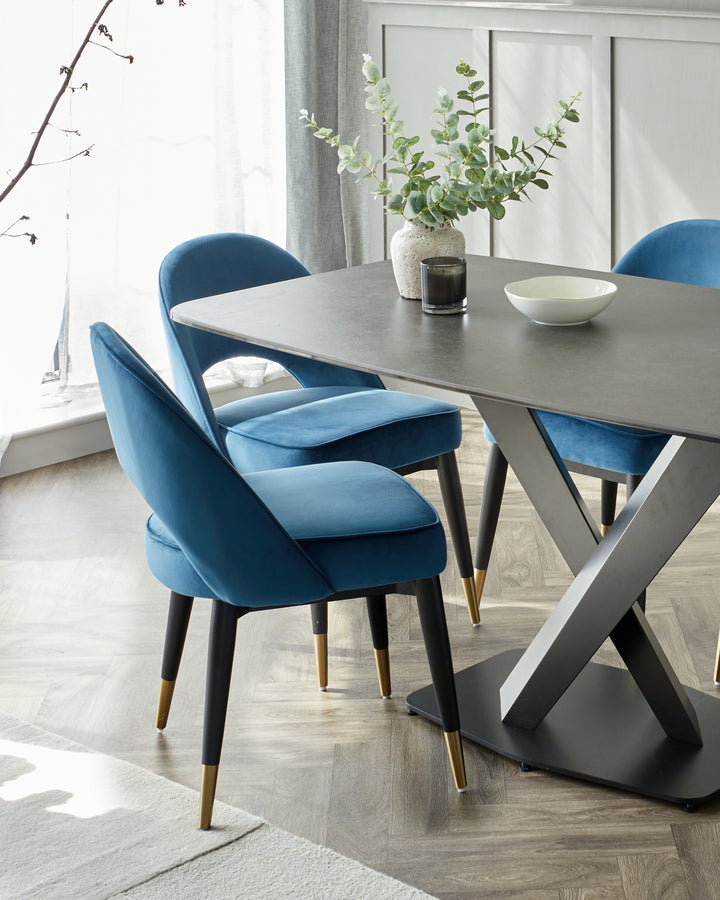 Modern dining set featuring a sleek dark grey table with a unique geometric base consisting of crossed metal legs, paired with four plush, velvet-upholstered chairs in a rich blue colour, accentuated with tapered black legs tipped with gold caps.