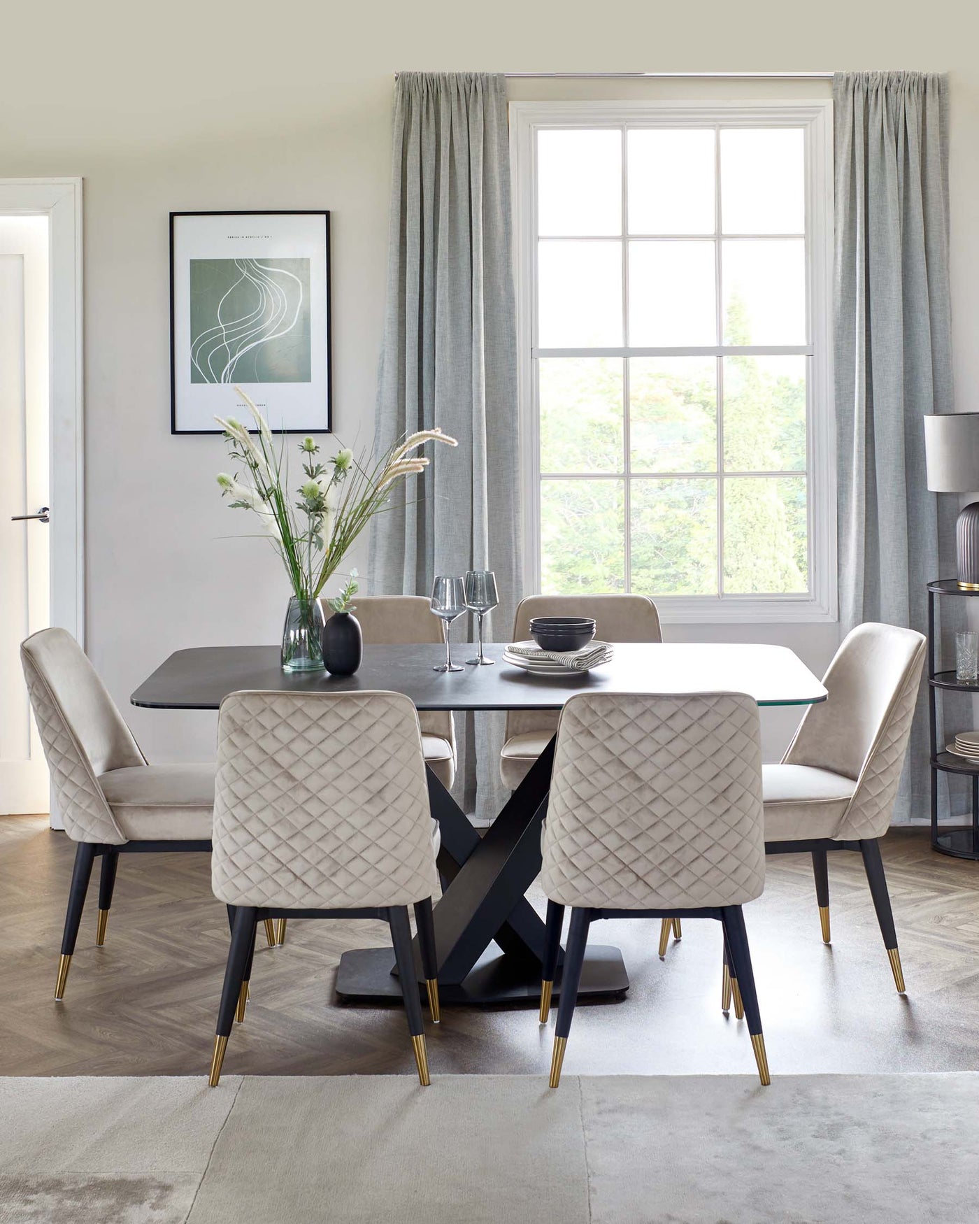 Elegant dining room featuring a modern rectangular table with a smooth dark top and a striking geometric black base, complemented by six plush, quilted dining chairs in a light taupe fabric. The chairs boast slender black legs with gold accent tips, matching the table's sophisticated aesthetic.