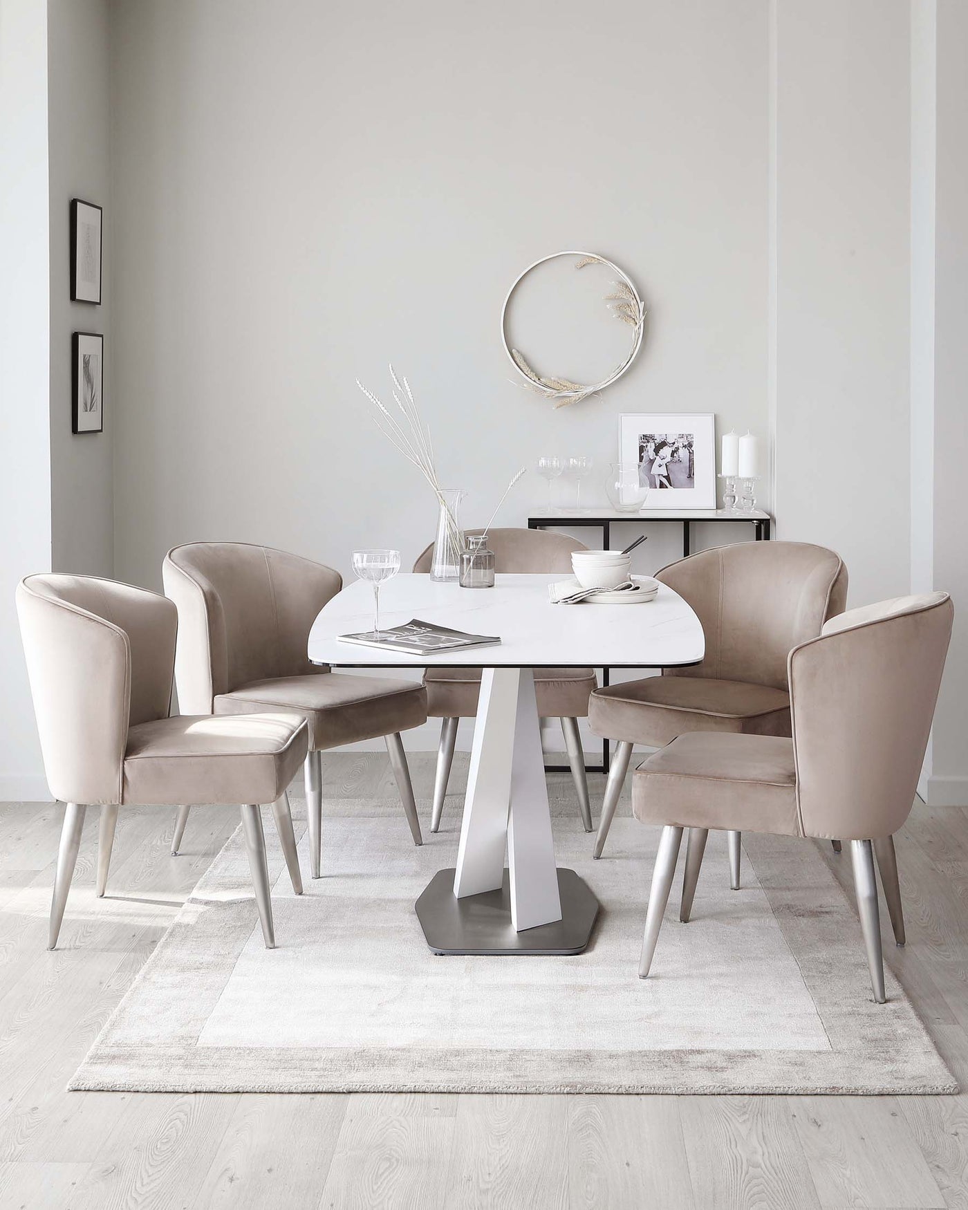 Modern dining room with a round white table featuring a geometric metallic base and a quartet of plush, taupe upholstered chairs with sleek tapered legs, set upon a textured light grey area rug.